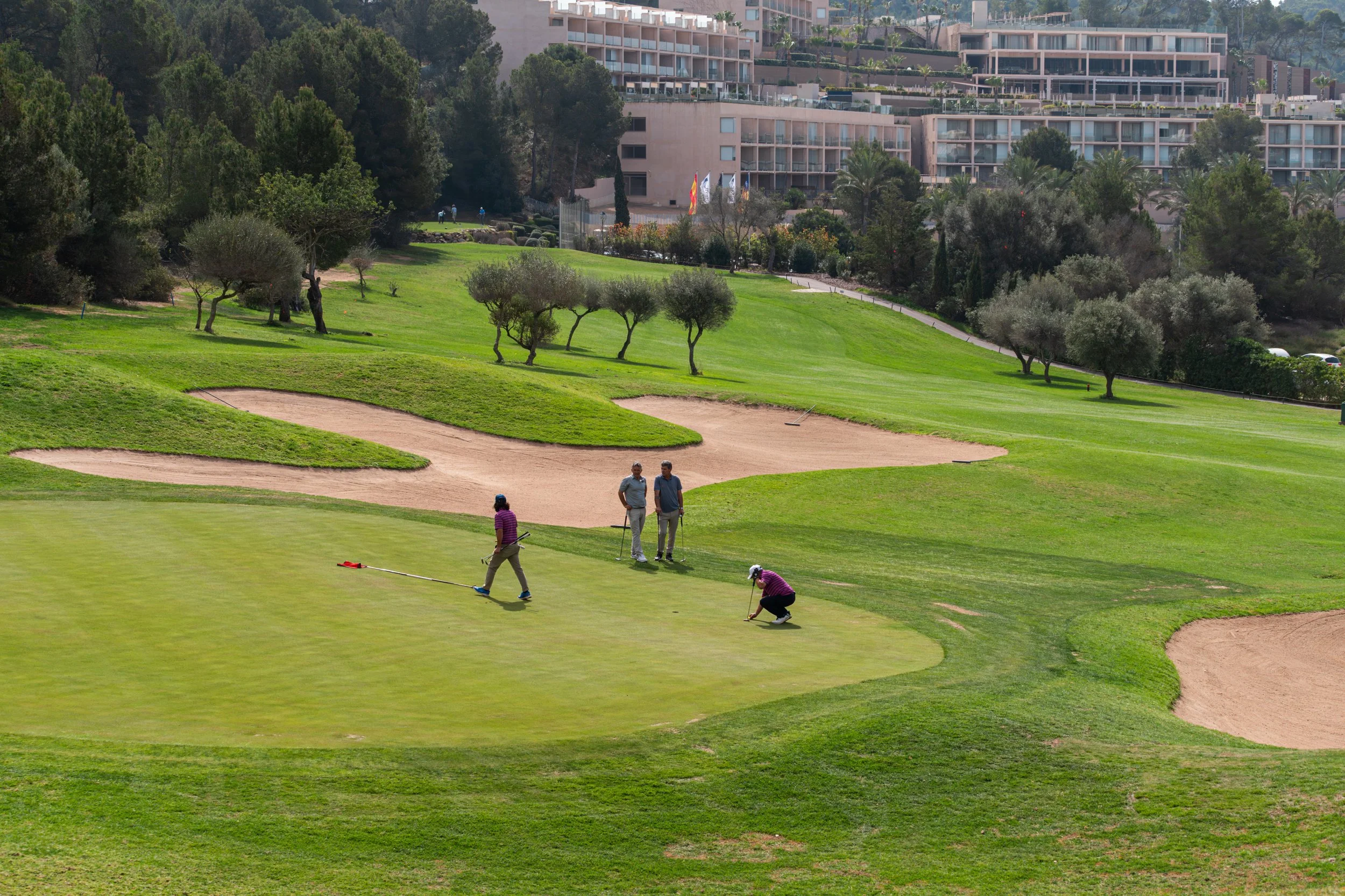 Personas jugando golf en un campo con áreas verdes, árboles y búnkeres, con edificios residenciales en el fondo.