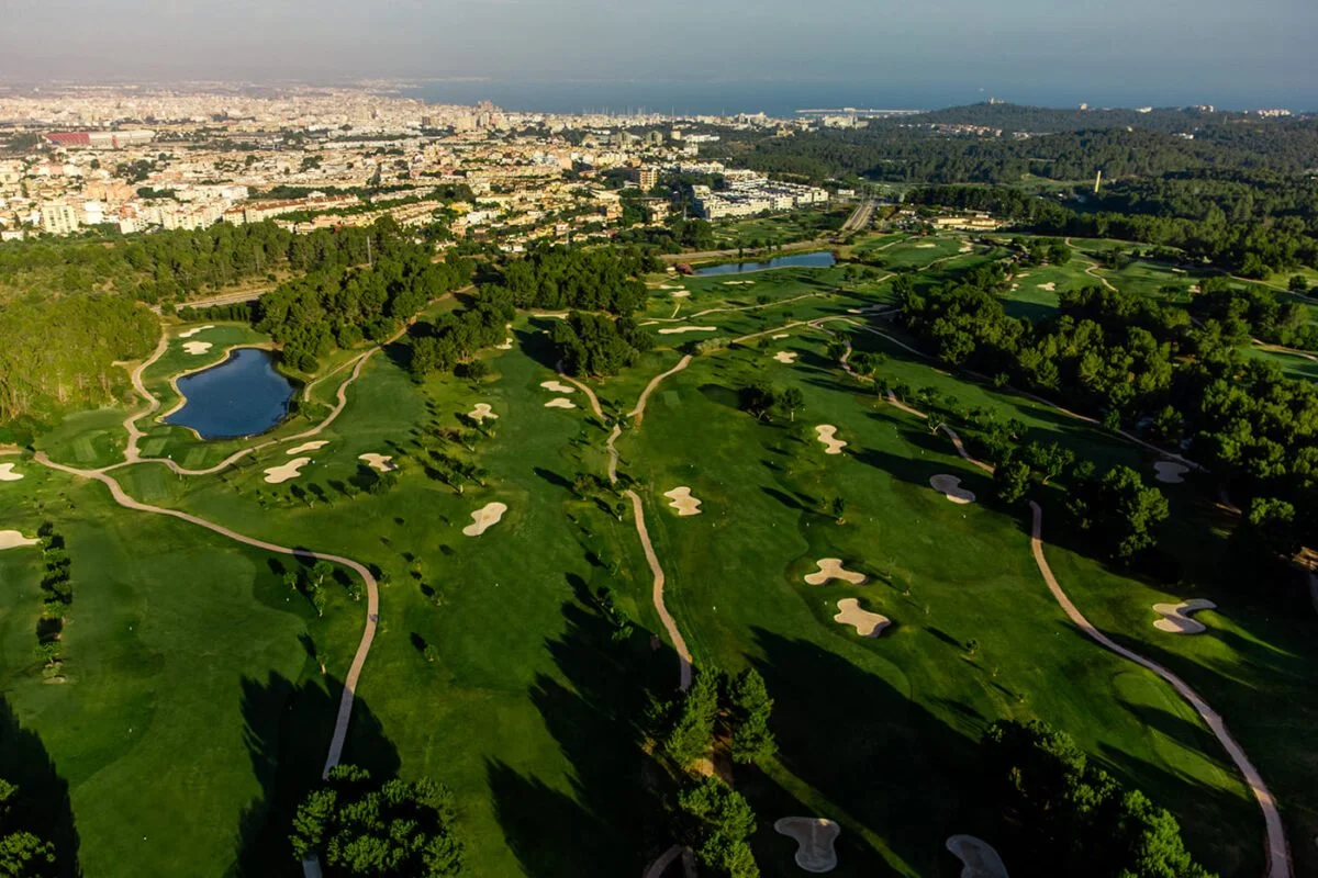 Vista aérea de un campo de golf con numerosos búnkers y lagos, rodeado de árboles y con una ciudad en el fondo.