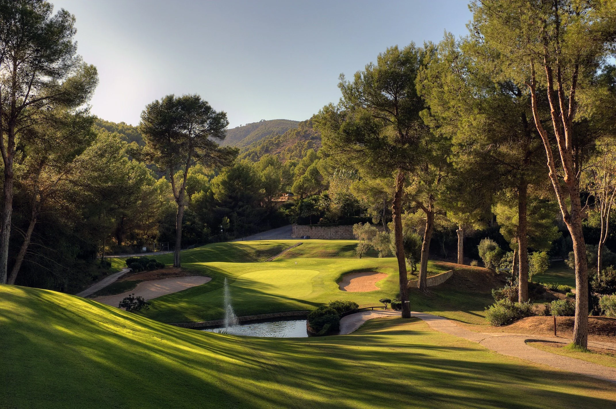 Campo de golf rodeado de árboles y montañas, con una fuente en el agua y caminos pavimentados.