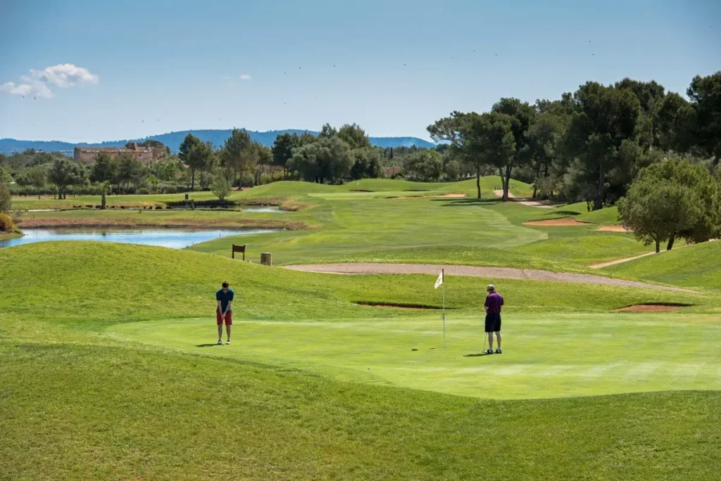 Dos personas jugandogolf en un campo de golf, con árboles, lagos y colinas verdes en un día soleado.