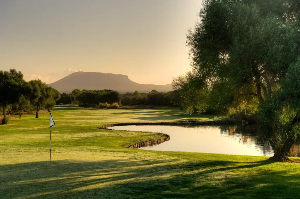 Un campo de golf con árboles, un pequeño lago y una bandera de golf, con montañas al fondo bajo un cielo despejado.