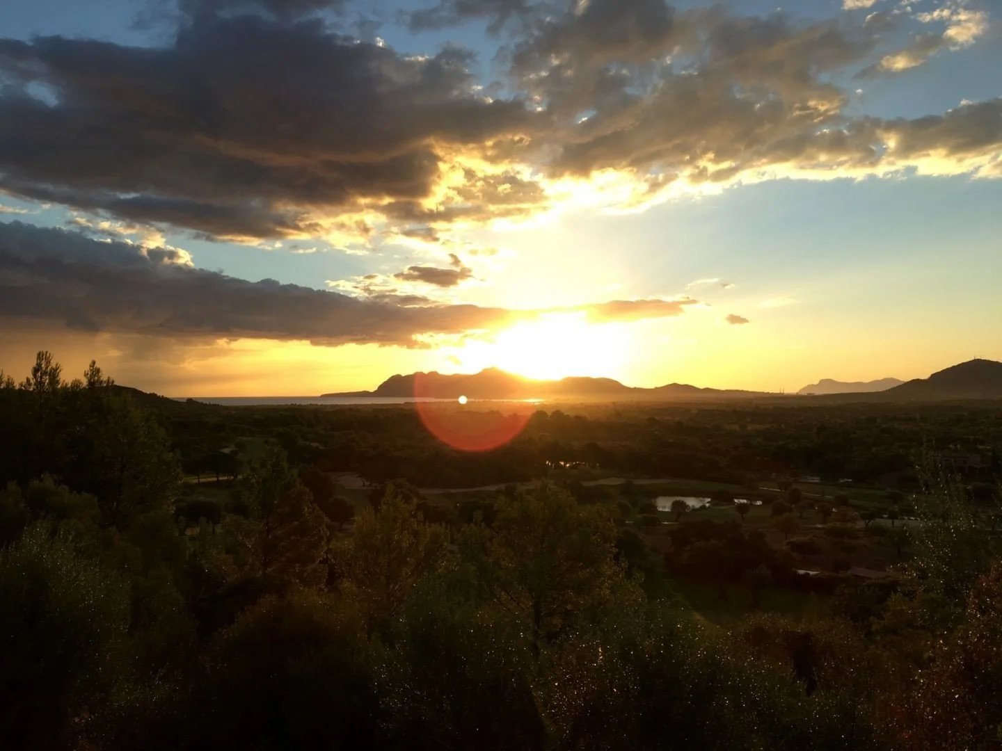 Atardecer sobre un paisaje con montañas y vegetación en primer plano, cielo con nubes oscuras y sol brillante