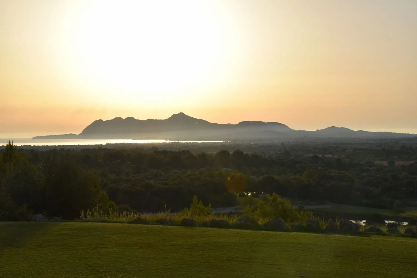 Paisaje con montañas al atardecer, árboles y césped en primer plano, reflejos en un cuerpo de agua.