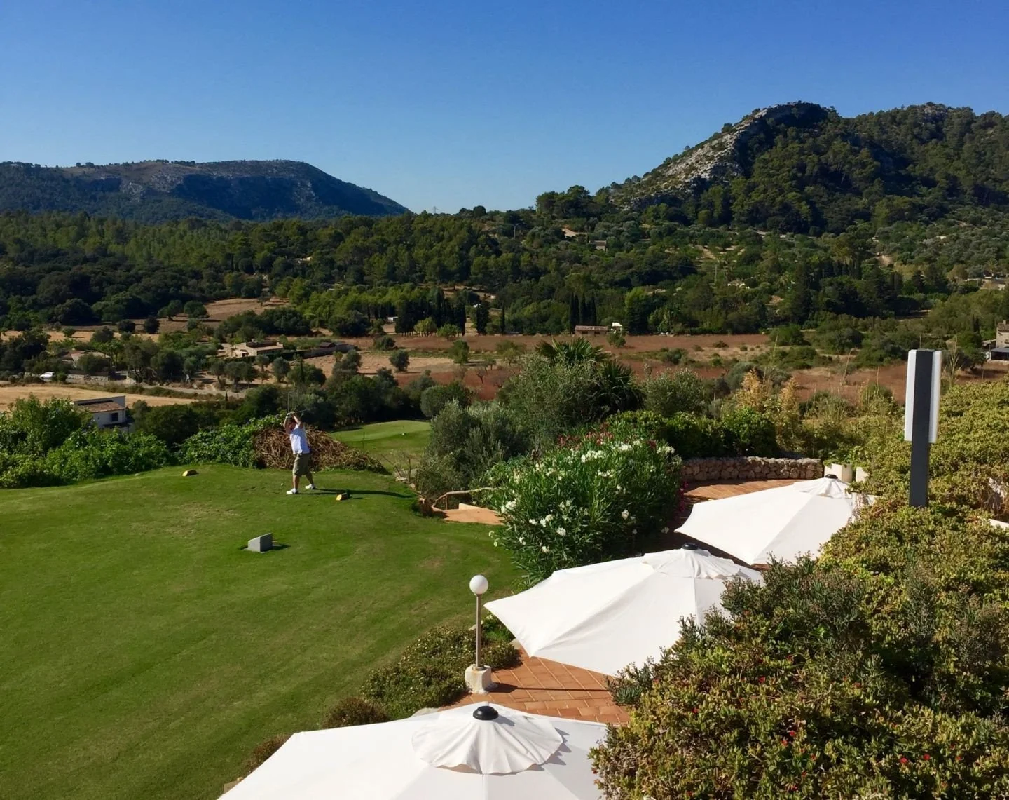 Persona jugando golf en un campo con montañas de fondo y sombrillas blancas en primer plano