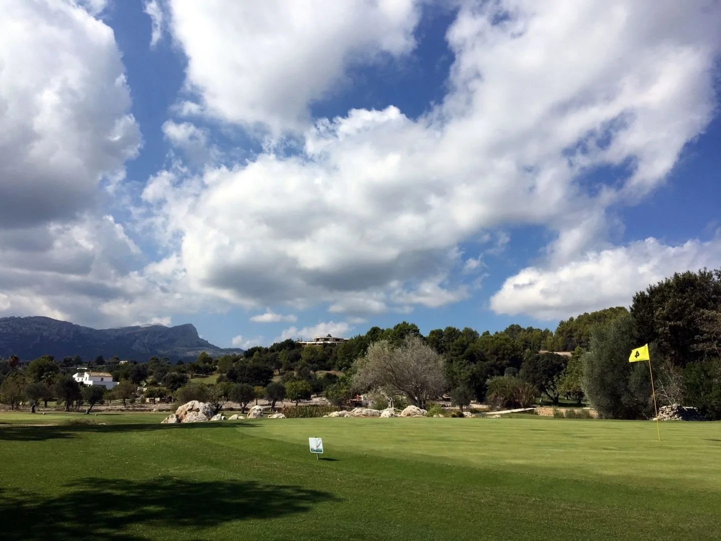 Campo de golf con bandera amarilla, césped bien cuidado, árboles dispersos y montañas al fondo bajo un cielo con nubes.