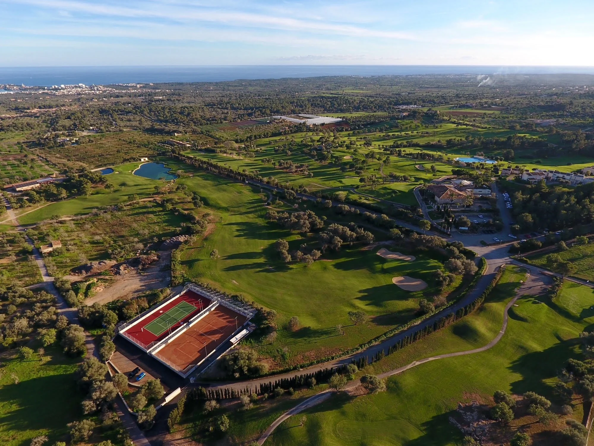Vista aérea de un campo de golf con áreas verdes, lagos, senderos y canchas de tenis, cerca de un área urbana y el océano en el fondo.