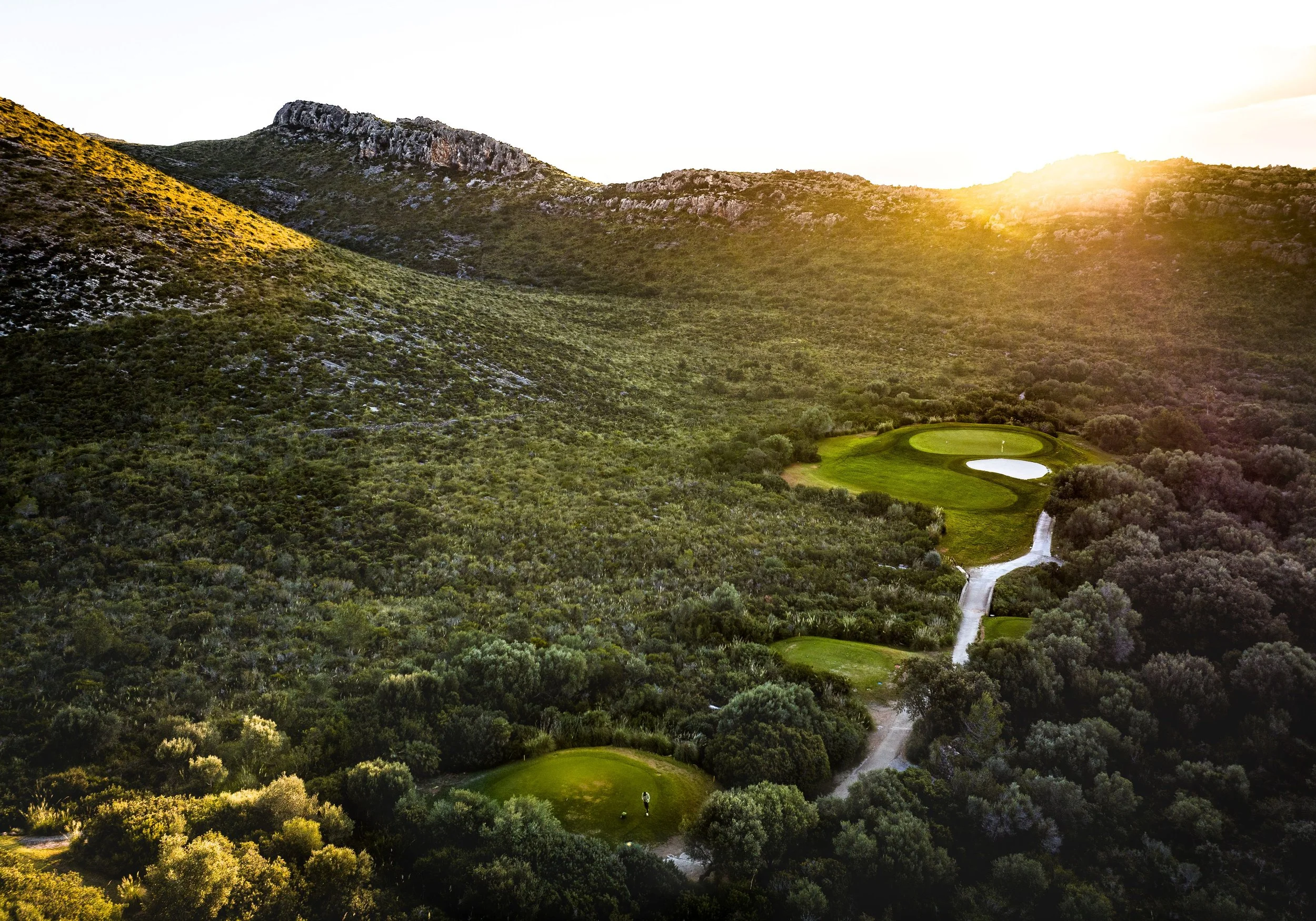 Campo de golf en un valle rodeado de colinas verdes y montañas, con la luz del sol al atardecer.