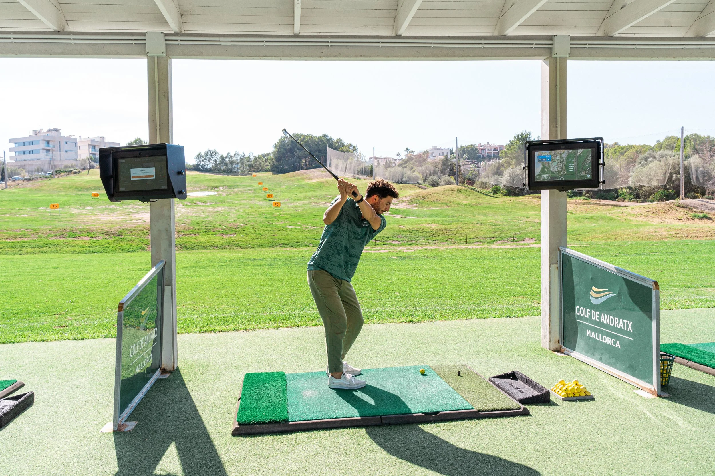 Un hombre en una zona cubierta de un campo de golf, practicando su swing con un palo, en un área cubierta en Mallorca, con pantallas y señalización del campo en el fondo.