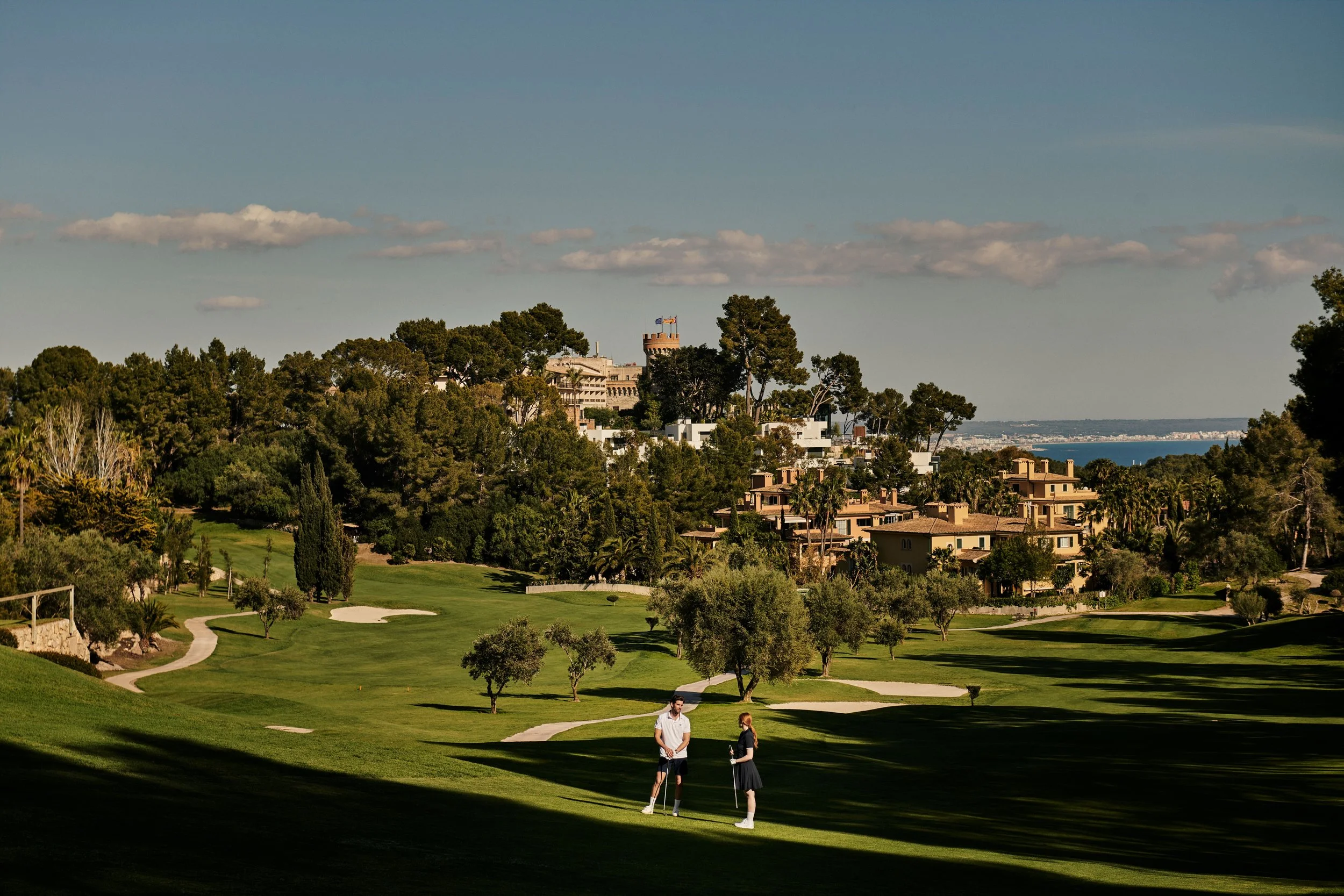 Vista panorámica de un campo de golf con dos personas conversando en primer plano, rodeado de árboles y casas residenciales, con una estructura en lo alto de una colina y vista al mar en el fondo.