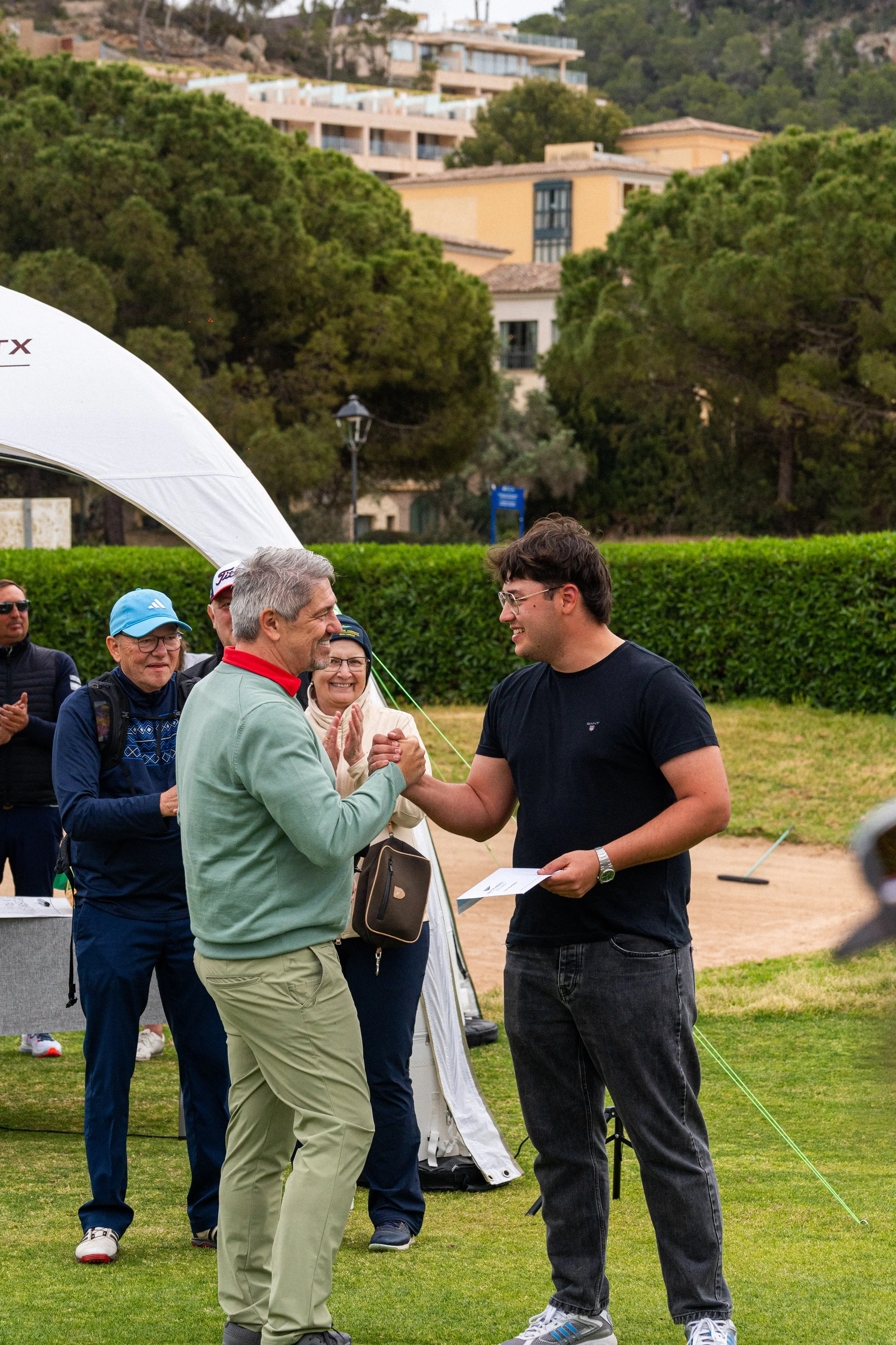 Dos hombres saludándose y dándose la mano en un campo de golf, con varias personas aplaudiendo al fondo y árboles y casas en el fondo.