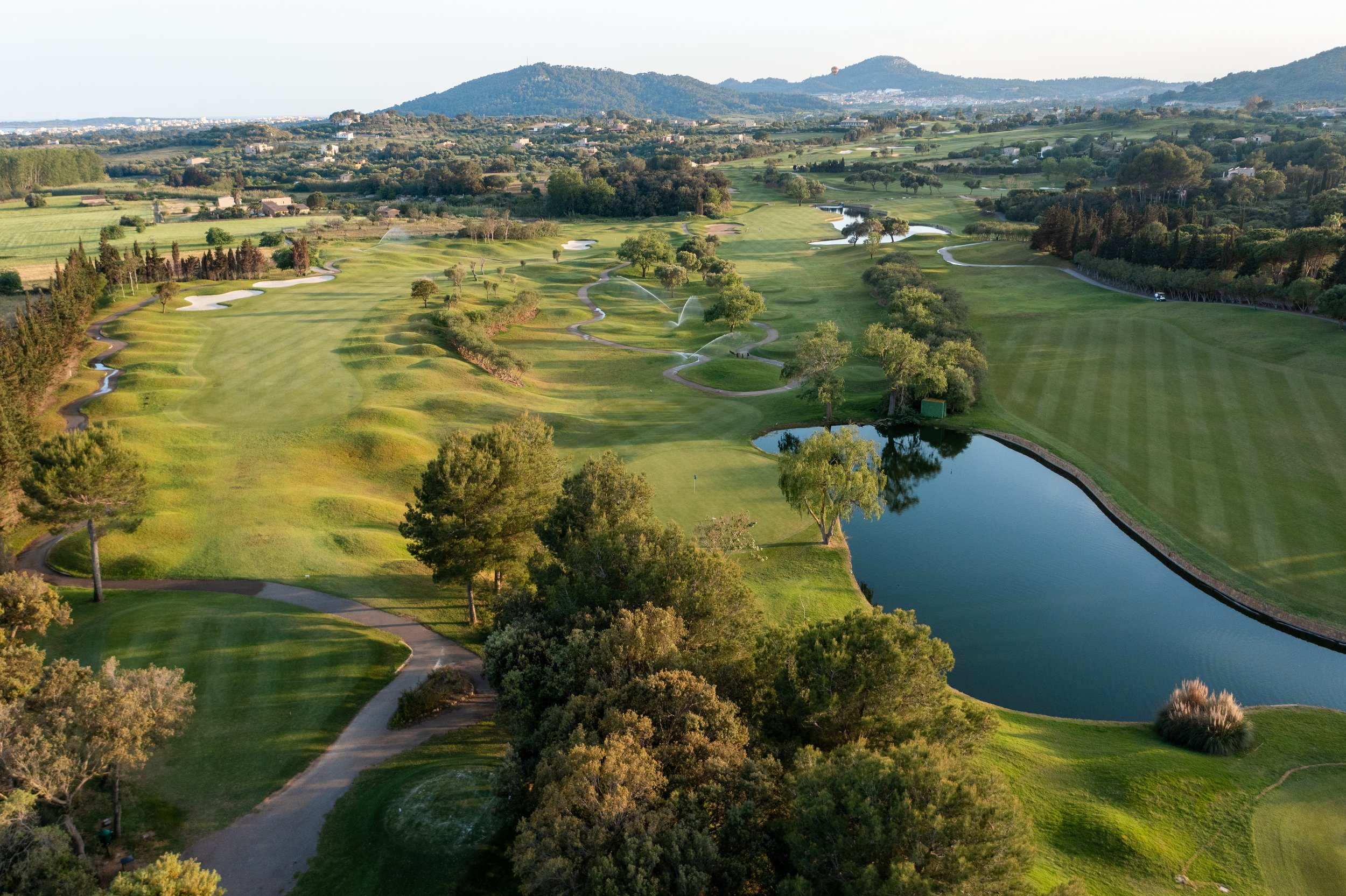 Vista aérea de un campo de golf con zonas verdes, lagos y árboles, rodeado de colinas y montañas en el fondo.