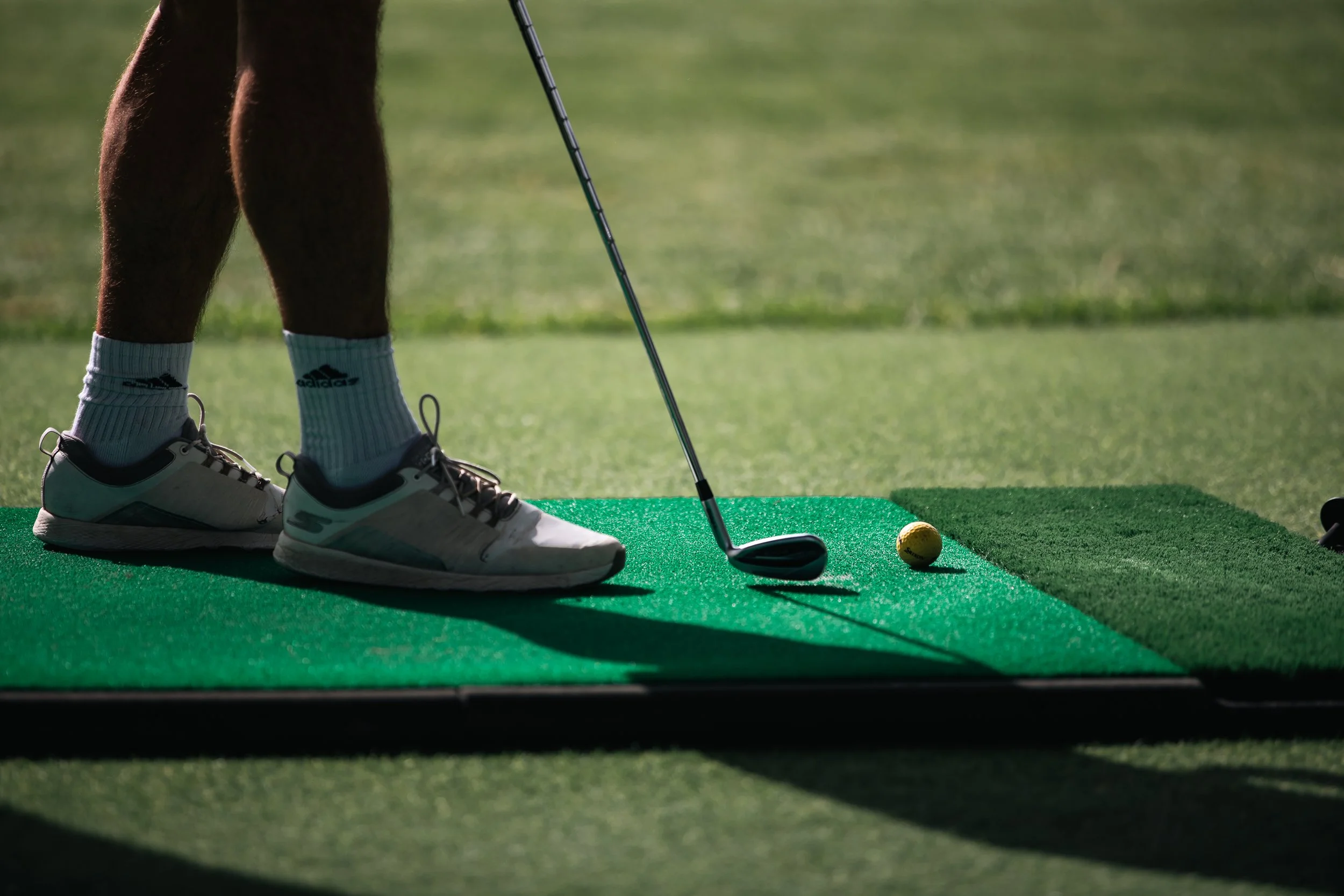 Persona en posición para jugar golf, con piernas, zapatos deportivos, y palo de golf en una alfombra verde con pelota de golf a un lado.