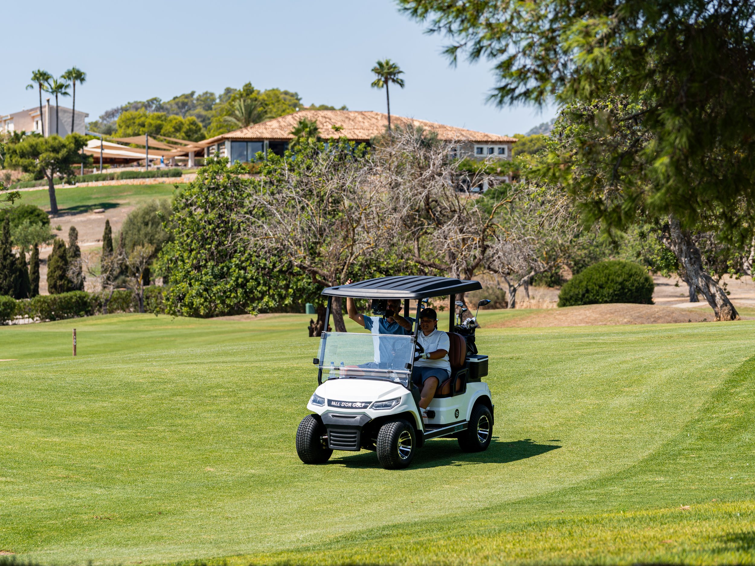 Dos personas en un carrito de golf en un campo de golf bien cuidado, con árboles y casas en el fondo.