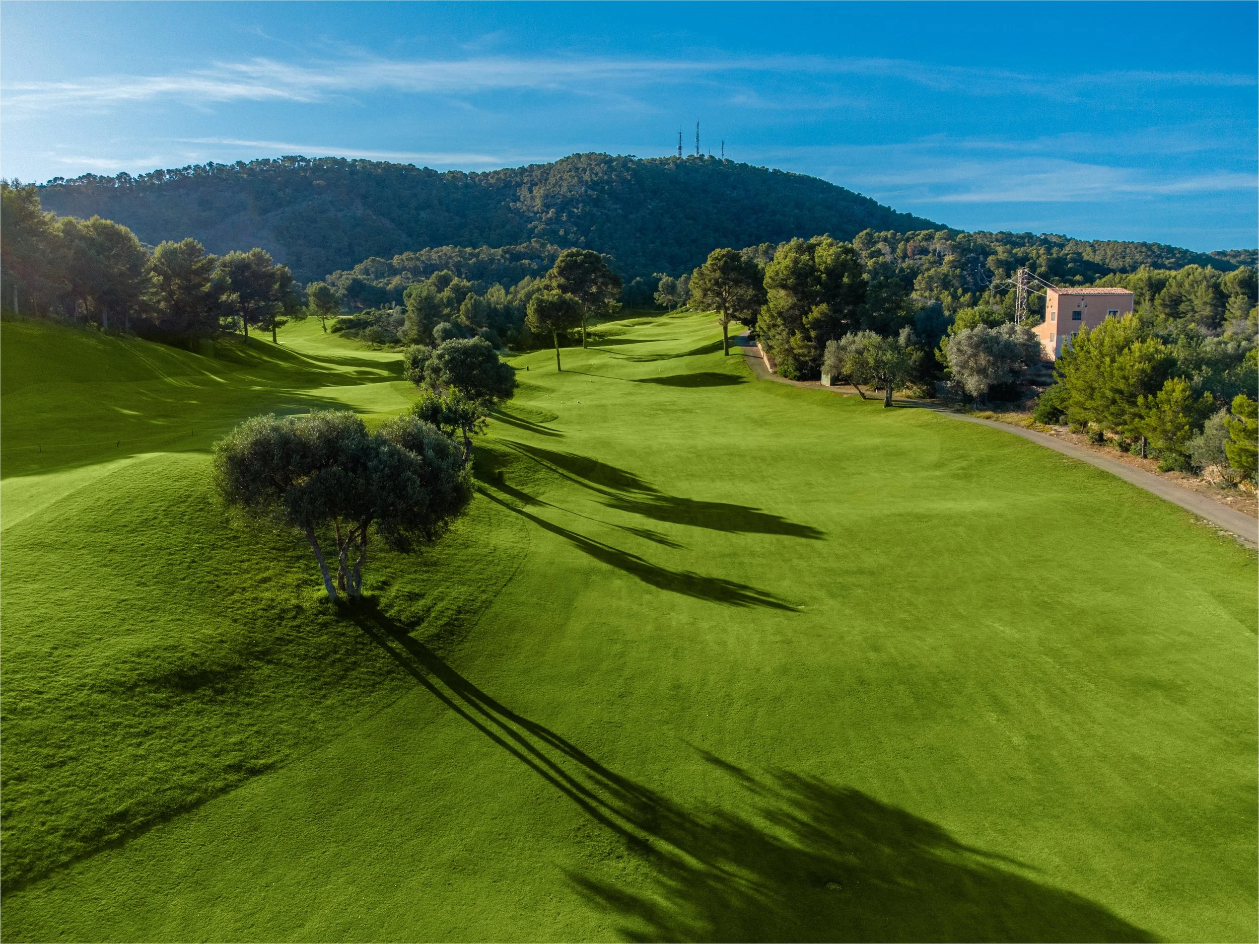 Campo de golf con árboles y colinas verdes, con una casa en el fondo y un cielo azul con nubes delgadas.