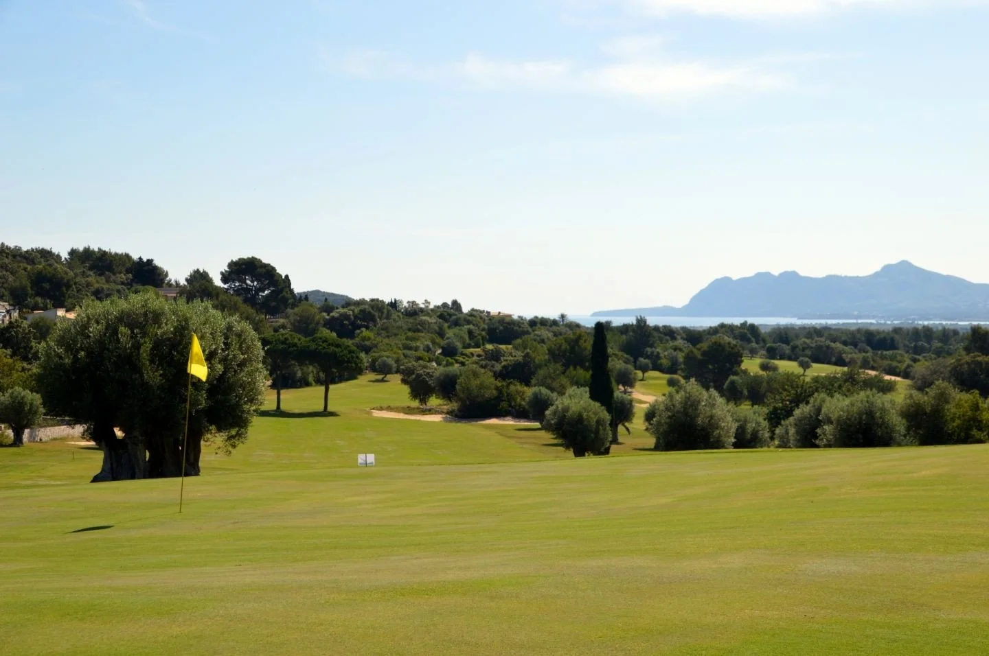 Vista de un campo de golf con césped bien cuidado, árboles dispersos y una bandera amarilla en primer plano, y en el fondo montañas y el mar bajo un cielo soleado.