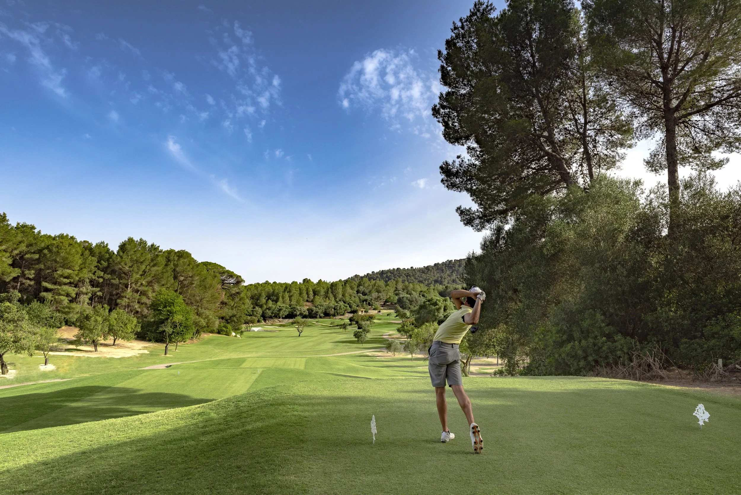 Jugador de golf en un campo de golf, preparándose para un golpe, rodeado de árboles y un cielo despejado.