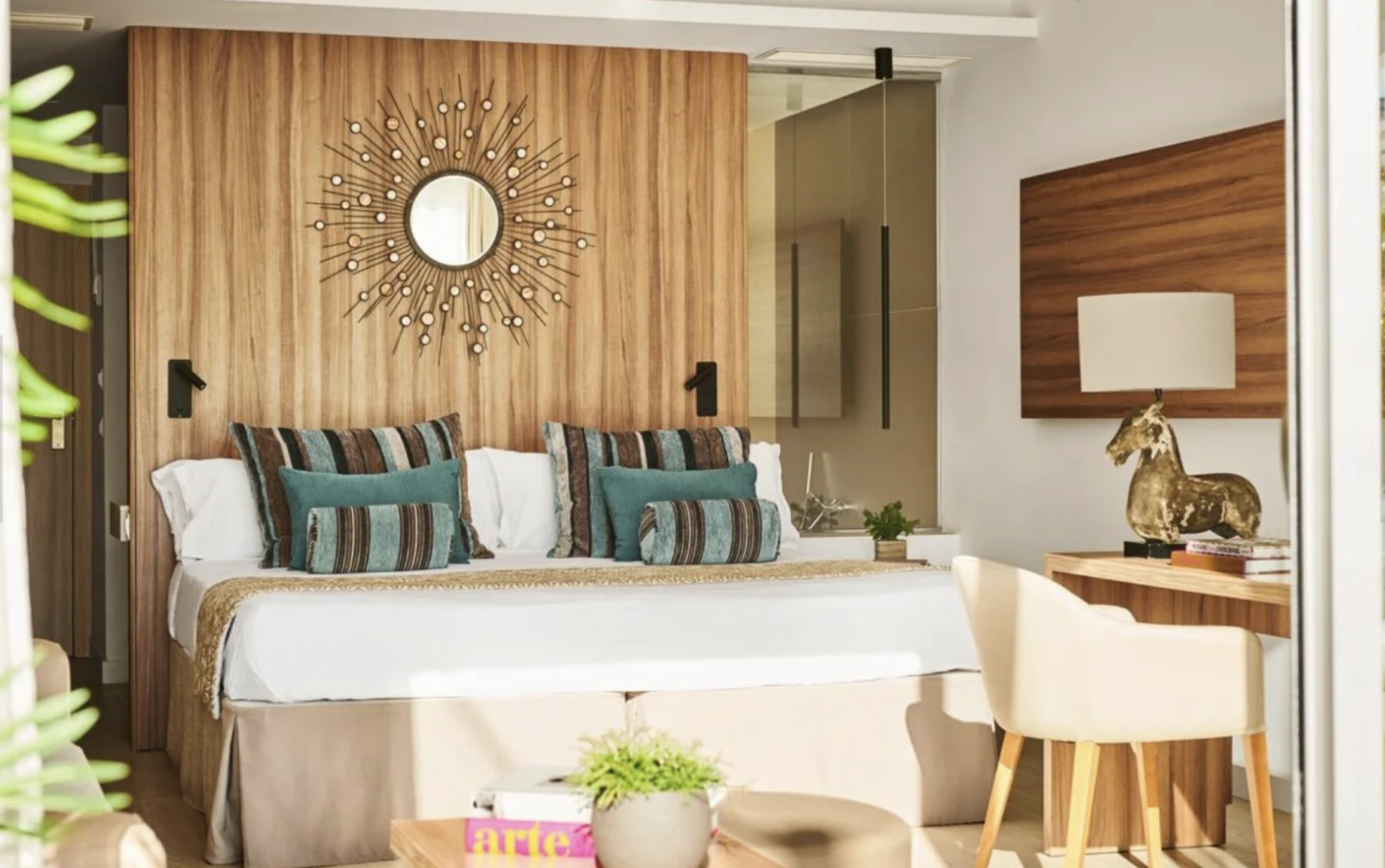 Recámara moderna con cabecera de madera, cortinas blancas, almohadas decorativas en tonos turquesa y marrón, espejo de sol con diseño de rayos, mesa de noche con lámpara, figura de caballo de madera, y sillón blanco con madera natural.