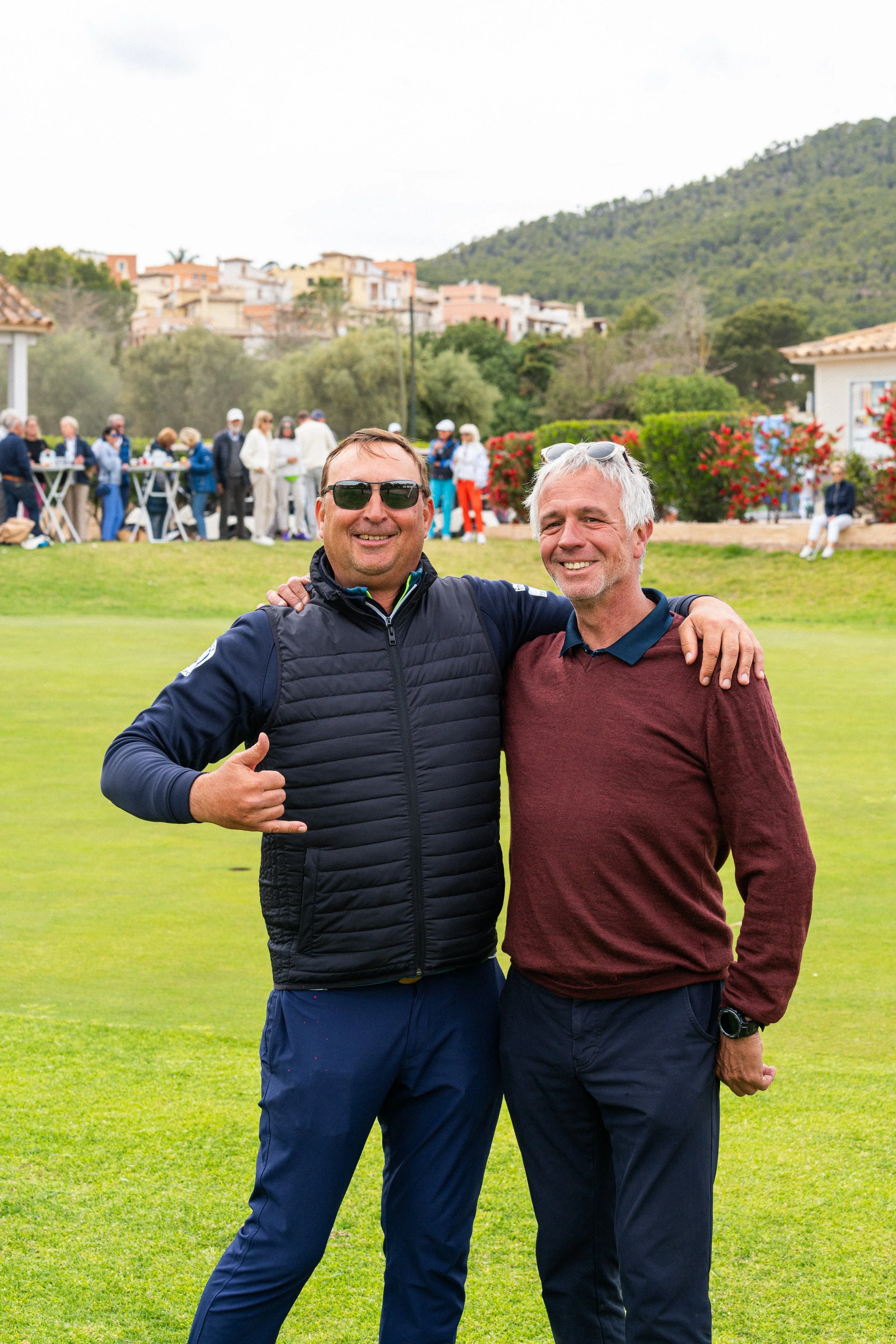 Dos hombres con ropa casual sonríen y se abrazan en un campo de golf, con otras personas en el fondo y casas con montañas en el horizonte.