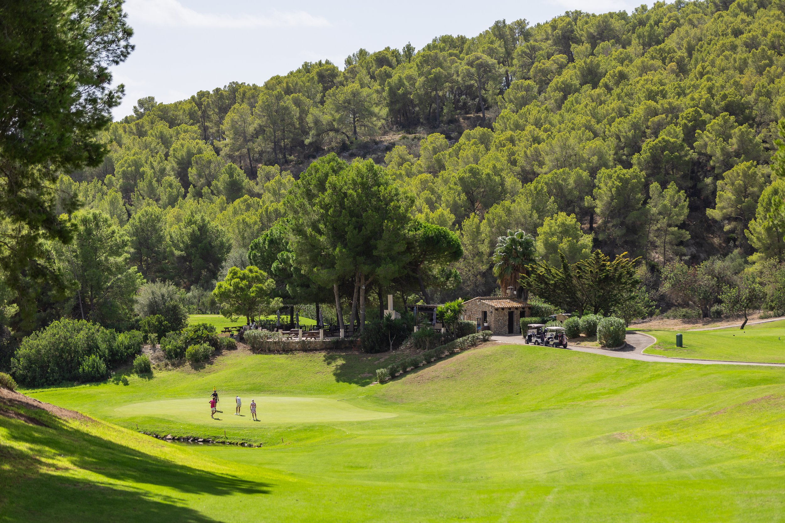 Campo de golf rodeado de árboles y colinas, con una casa de estilo rústico y unos carros de golf, y personas jugando al golf en el césped.