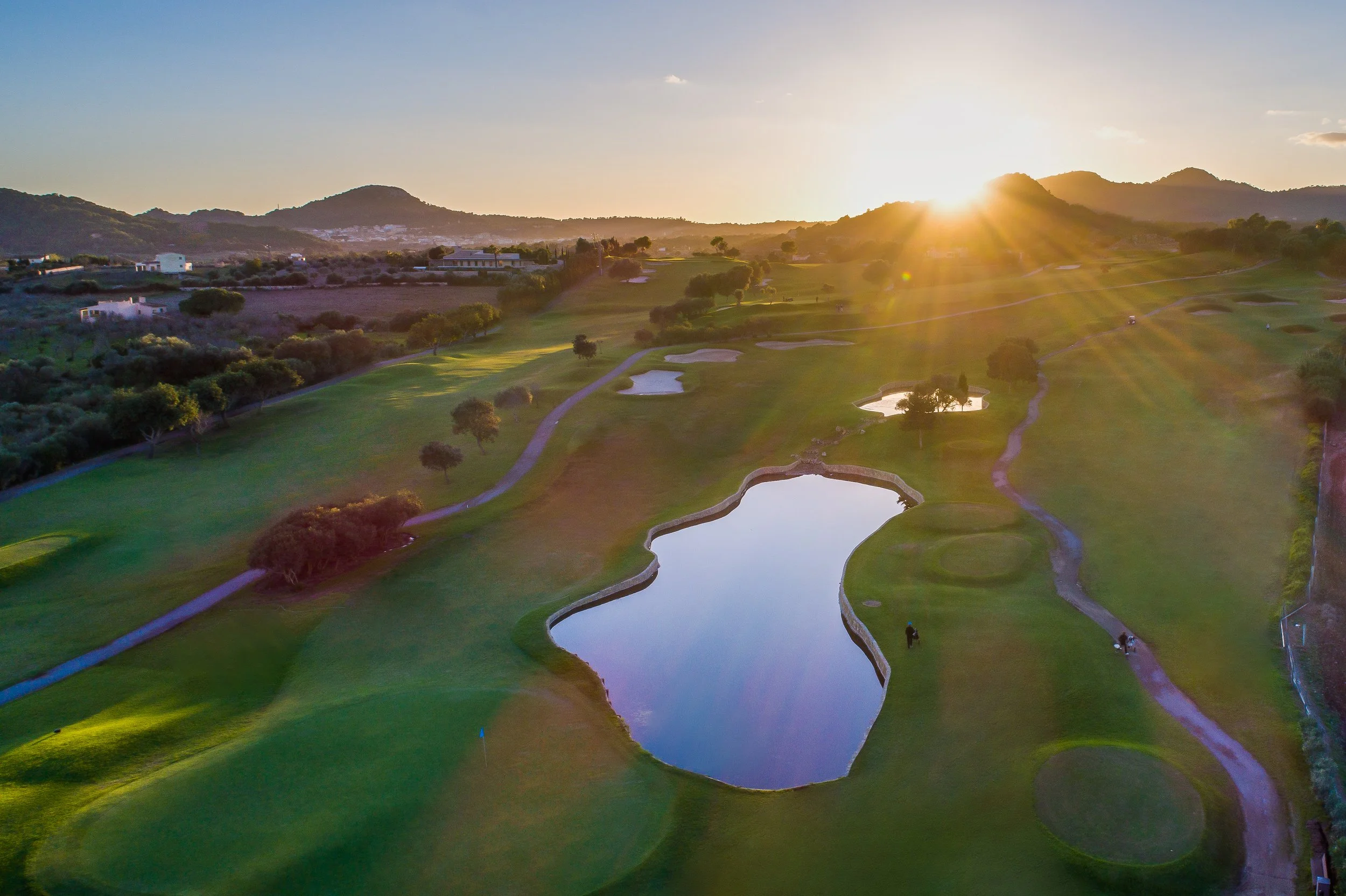 Paisaje de un campo de golf con estanques y árboles al atardecer, con montañas y casas en el fondo.