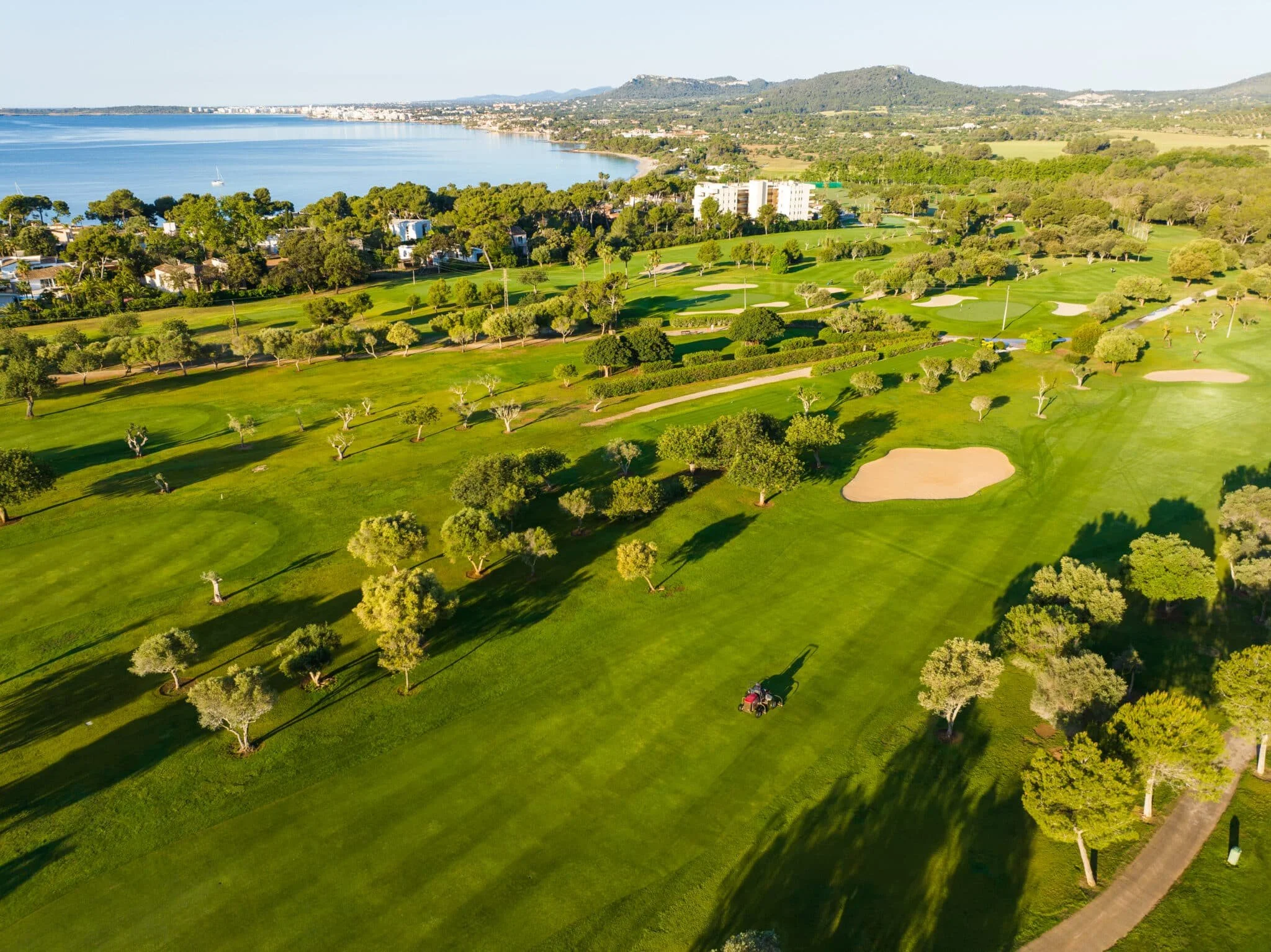 Campo de golf con árboles, arena y un carrito de golf, cerca de un cuerpo de agua y áreas urbanas en el fondo.