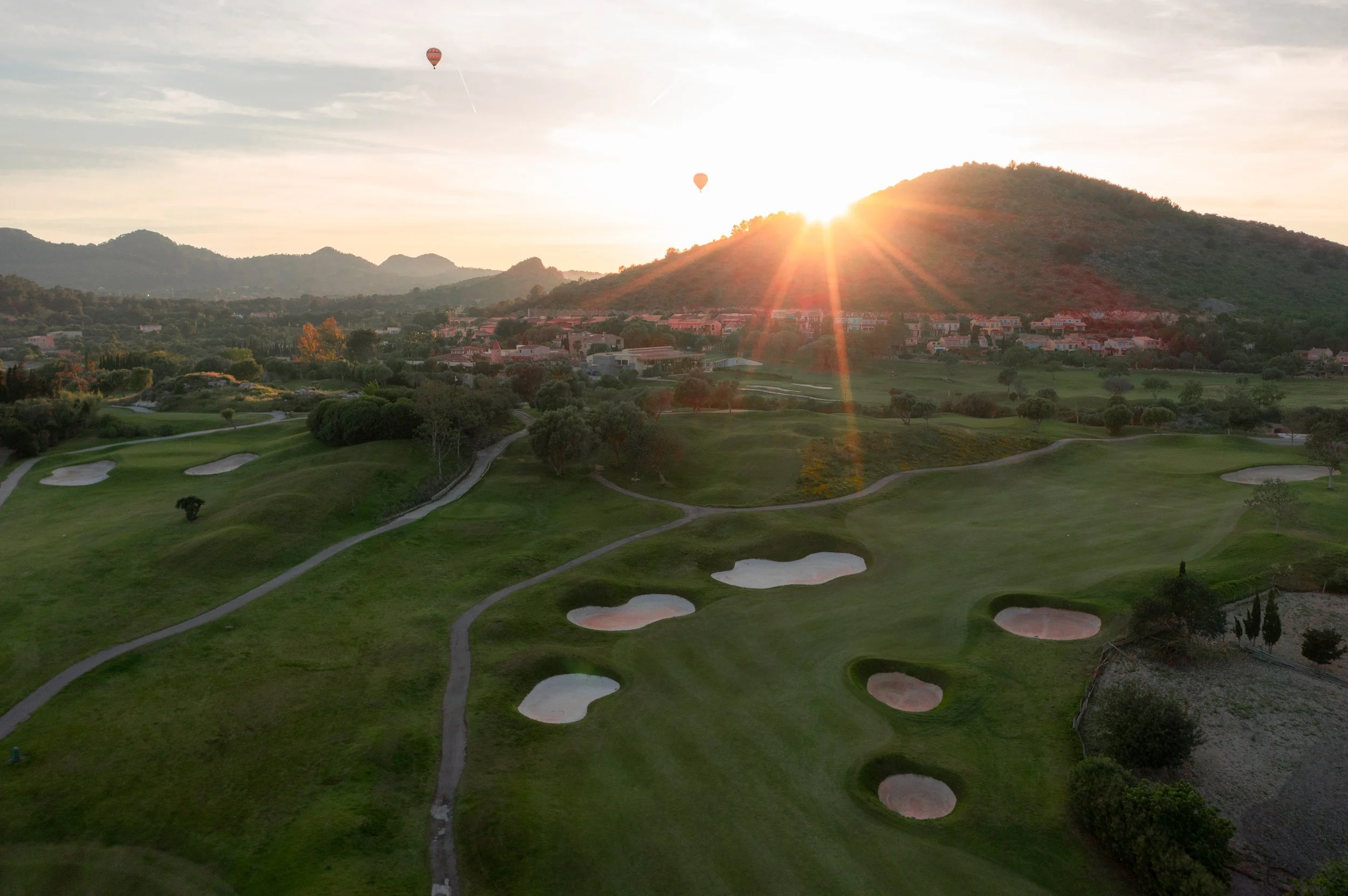 Vista aérea de un campo de golf al atardecer, con colinas y casas en el fondo, y globos aerostáticos en el cielo
