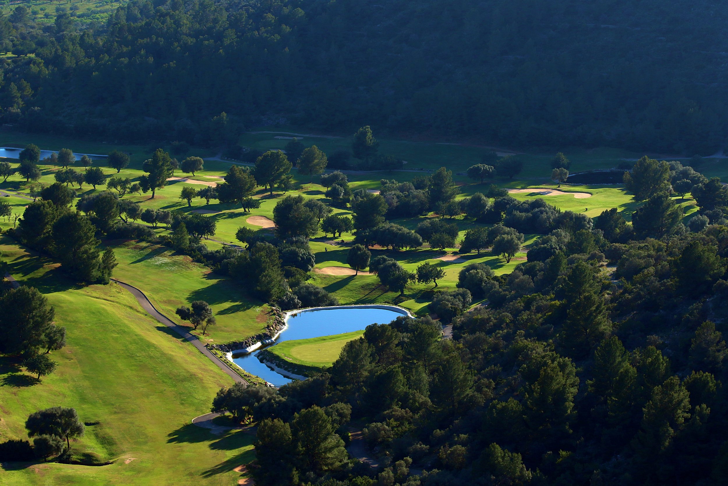 Vista aérea de un campo de golf rodeado de árboles y colinas, con un pequeño lago y caminos