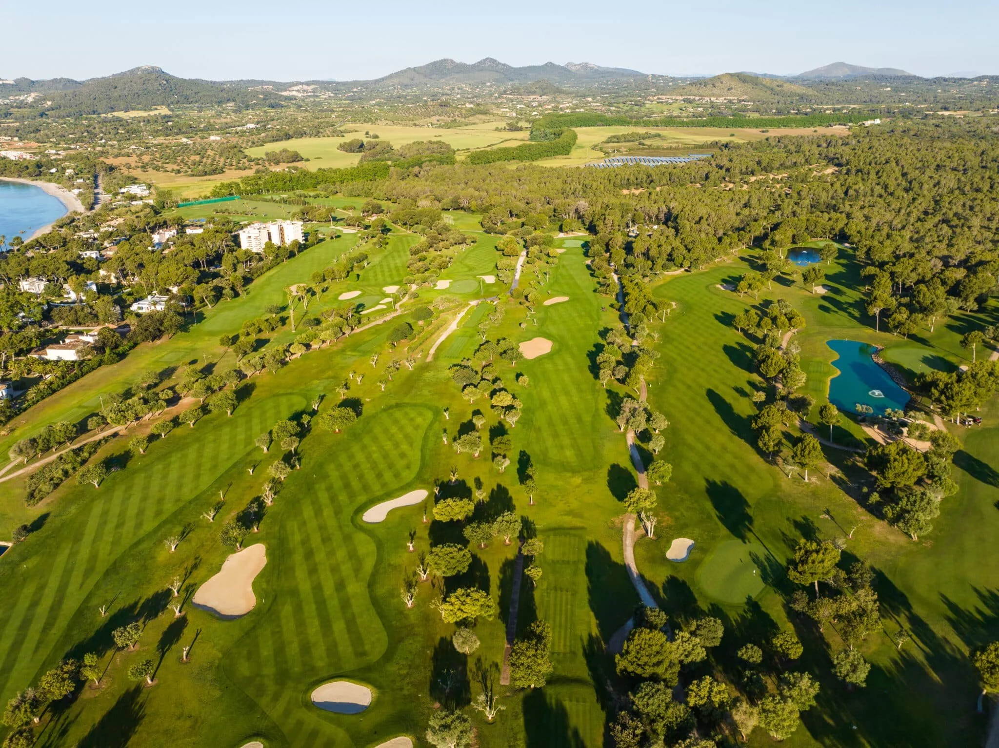 Vista aérea de un campo de golf con áreas verdes, árboles, lagunas y caminos pavimentados, en un entorno natural con montañas al fondo.
