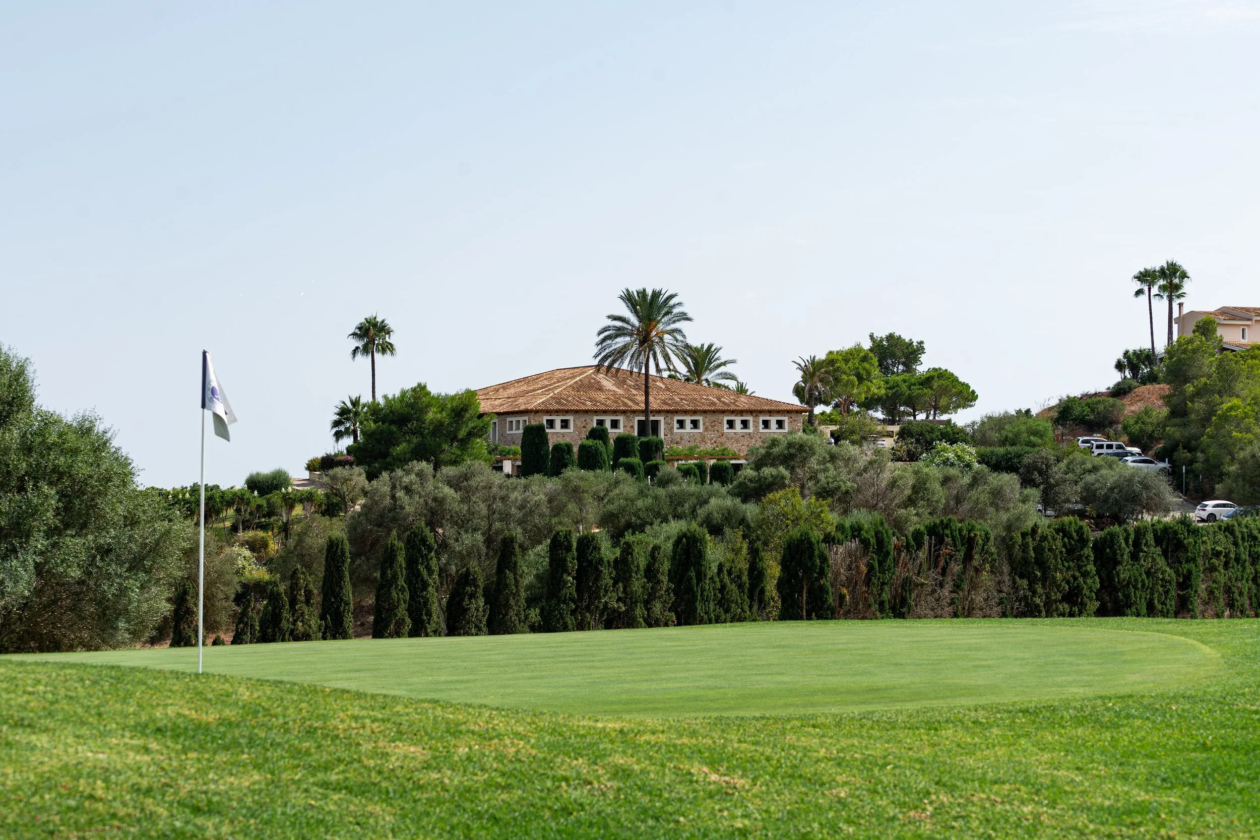 Campo de golf con bandera y casa en una colina rodeada de árboles y palmeras.