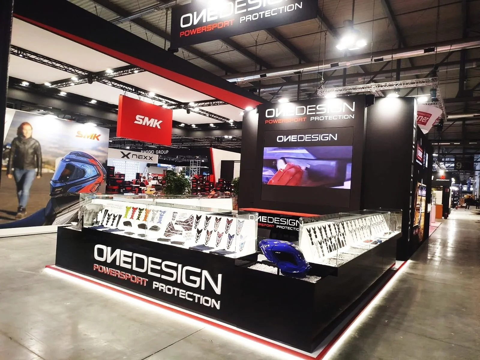 Stand espositivo di OVEDESIGN con caschi da moto e protezioni, con large schermo e logo in evidenza, al centro di una fiera di settore.