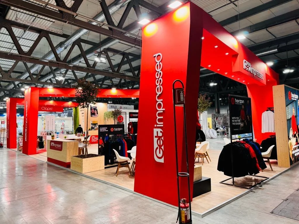 Stand di abbigliamento con struttura rossa, presenti vestiti in esposizione, sedie bianche e alcuni clienti all'interno, in un centro commerciale o fiera.