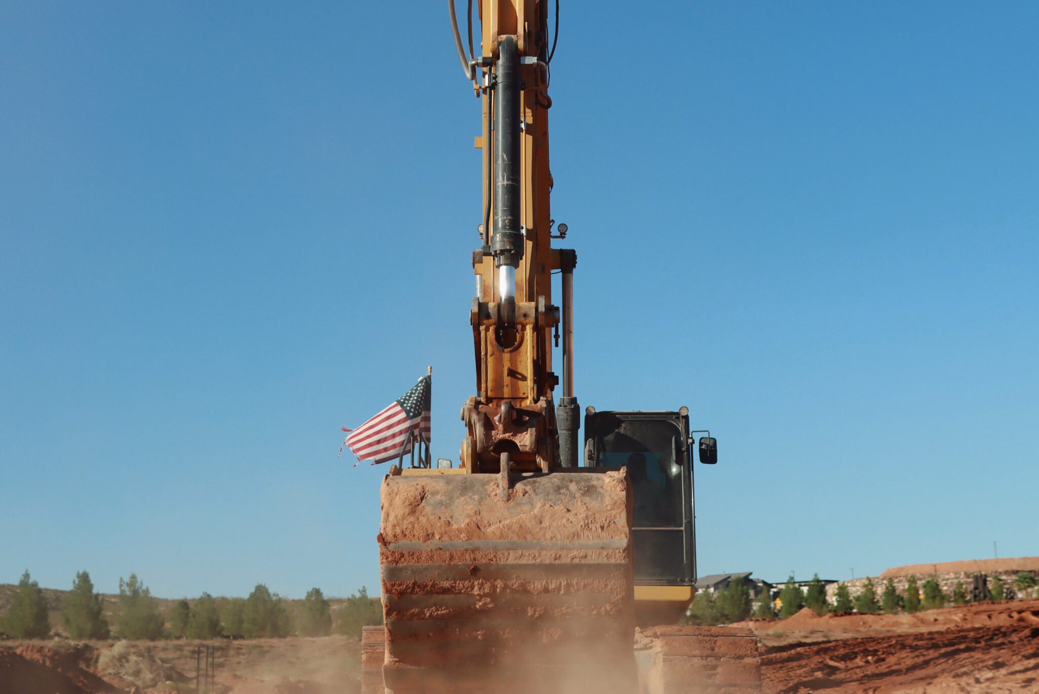 A construction excavator is digging into the red dirt at a construction site with an American flag waving in the background under a clear blue sky.