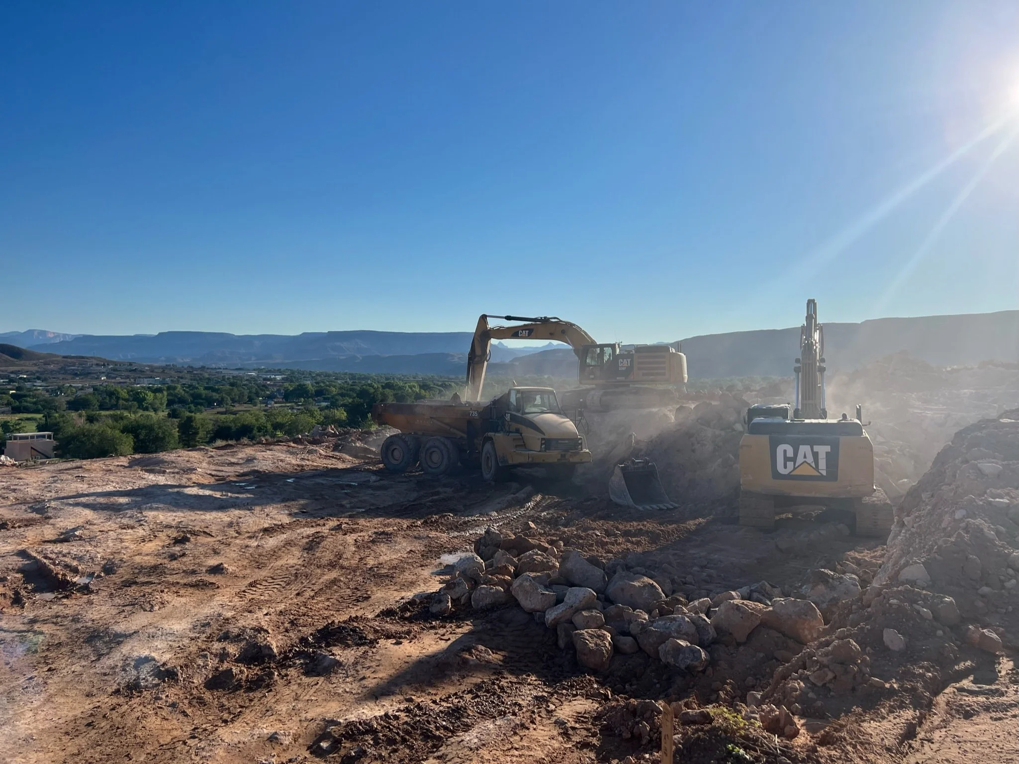 Construction site with excavators and a dump truck working on land, with mountains and greenery in the background and the sun shining brightly in the sky.
