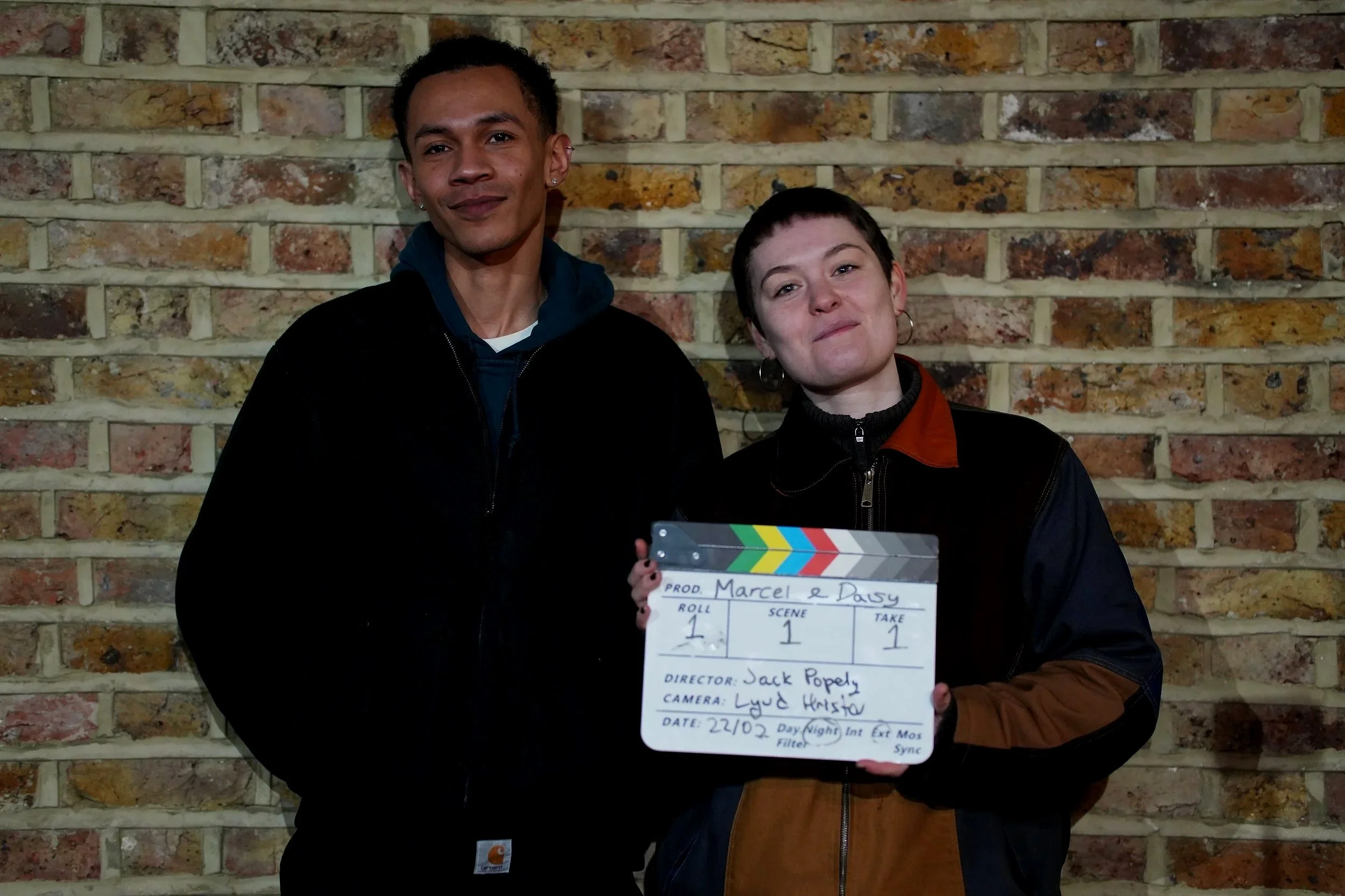Two people standing in front of a brick wall, having just wrapped their showreel, one holding a film clapper board
