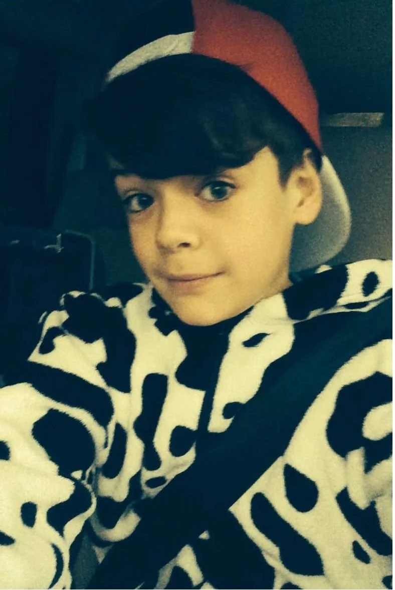 A young boy wearing a cow-patterned hoodie and a red and gray baseball cap, sitting in a car.
