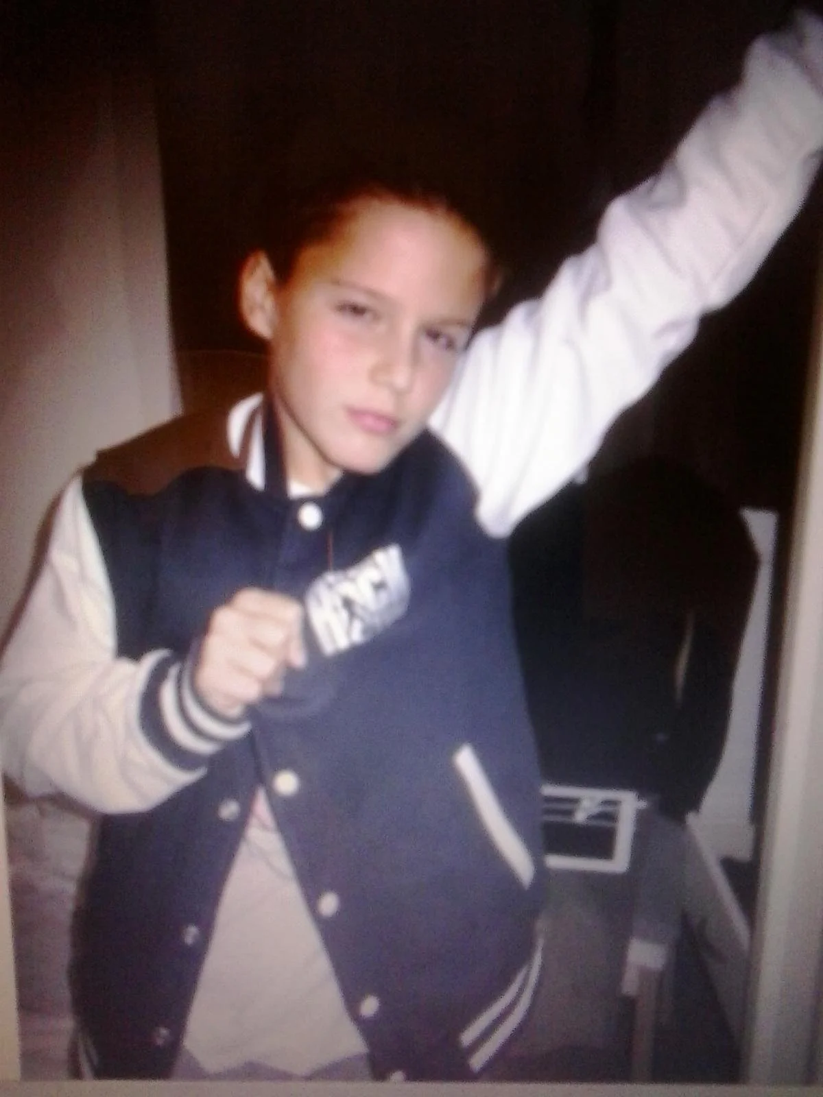 Young boy with short dark hair, wearing a black and white varsity jacket with a sweatshirt underneath, posing with one arm raised and a fist near his chest.