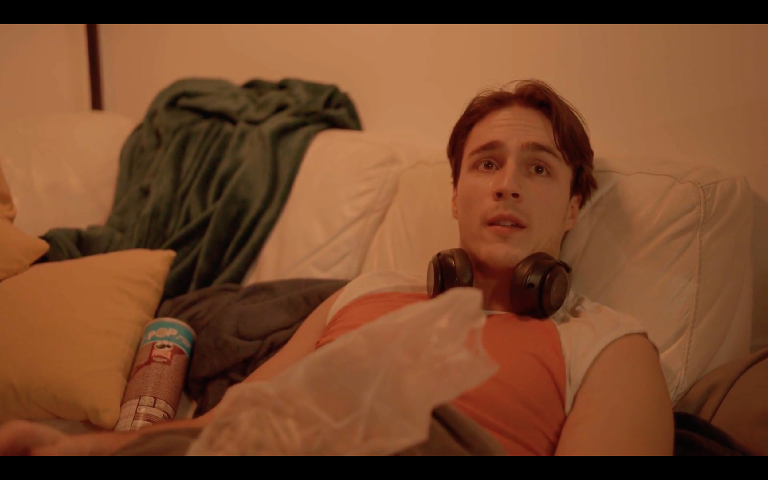 A young man with short brown hair lies on a bed, wearing orange and white clothing with headphones around his neck. He has a surprised or concerned expression. Behind him are pillows and a blanket.