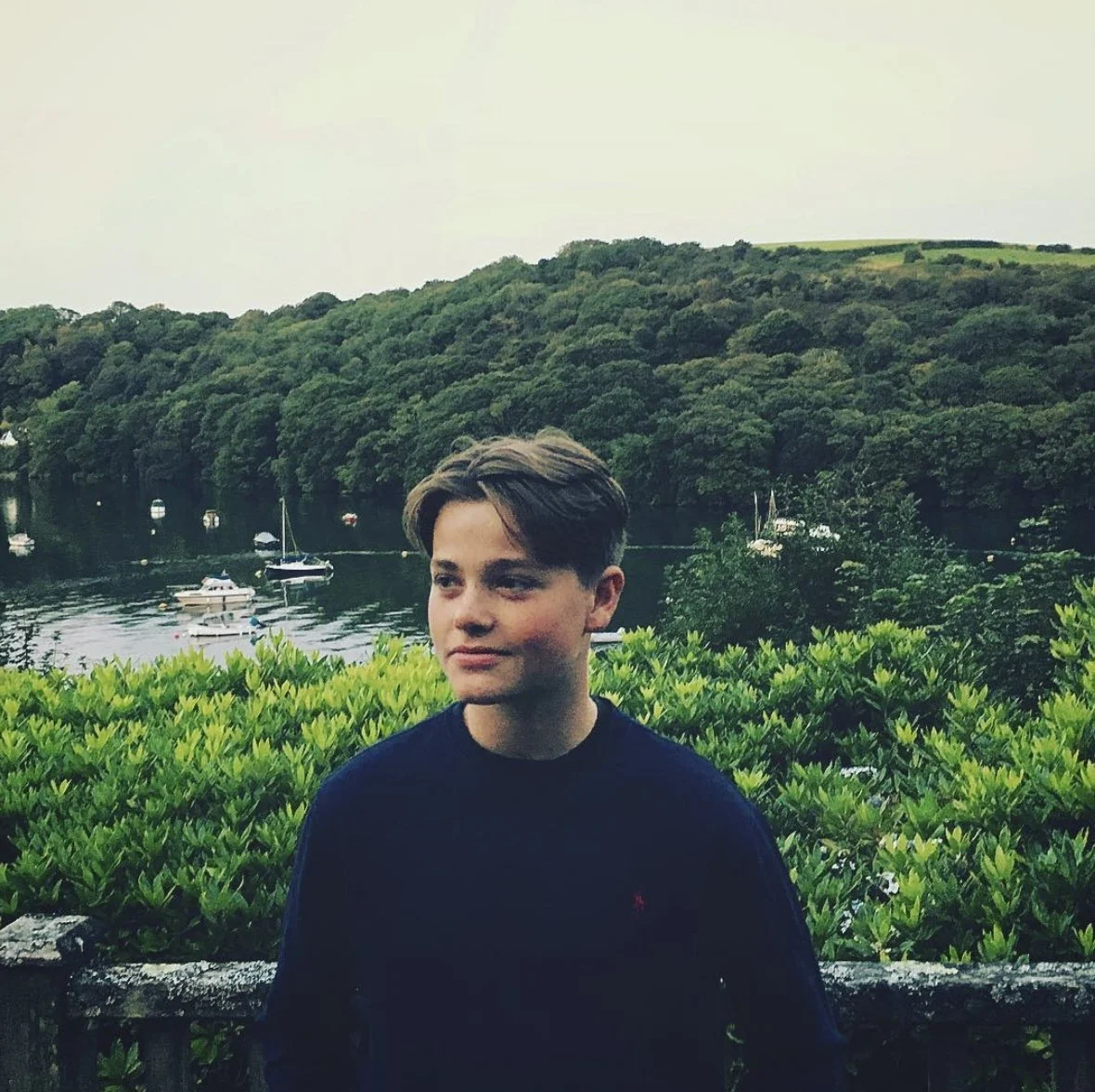 A young man with brown hair standing outdoors near a stone wall with greenery, overlooking a waterfront with boats and a wooded hill in the background.