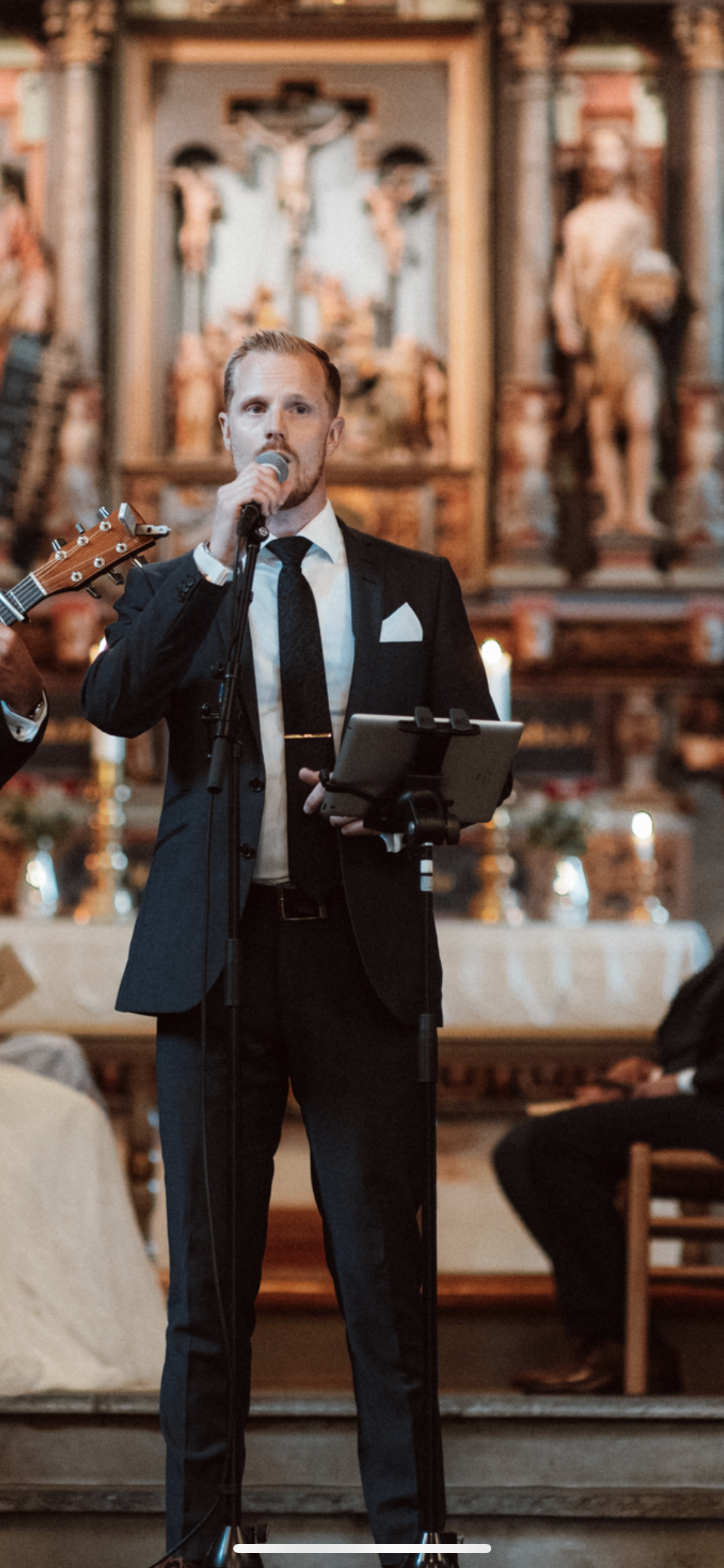 En mann i sort dress og slips holder mikrofon og taler i en kirke, med alter og religiøse figurer i bakgrunnen.