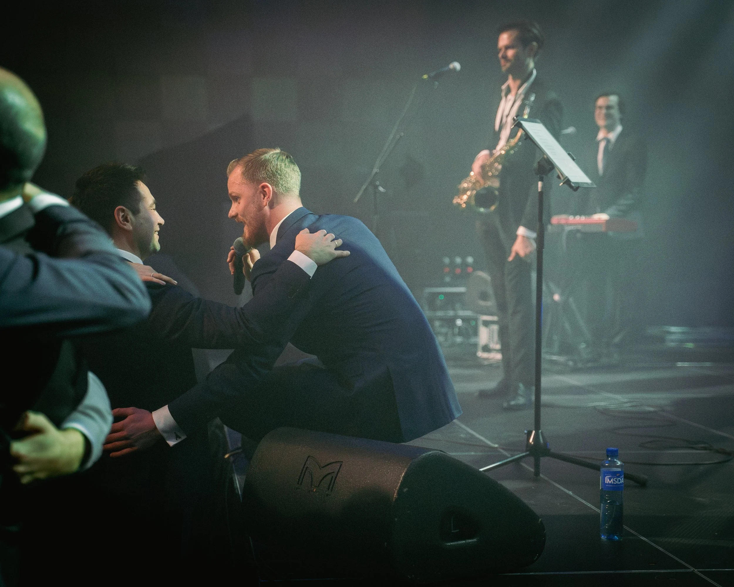 Tre menn i forretningskostymer som smiler og har en samtale ved en konsert eller event. En person spiller sakte på saxofon i bakgrunnen, mens en annen står ved en mikrofon. Scenen er svakt opplyst og det er musikkutstyr rundt.