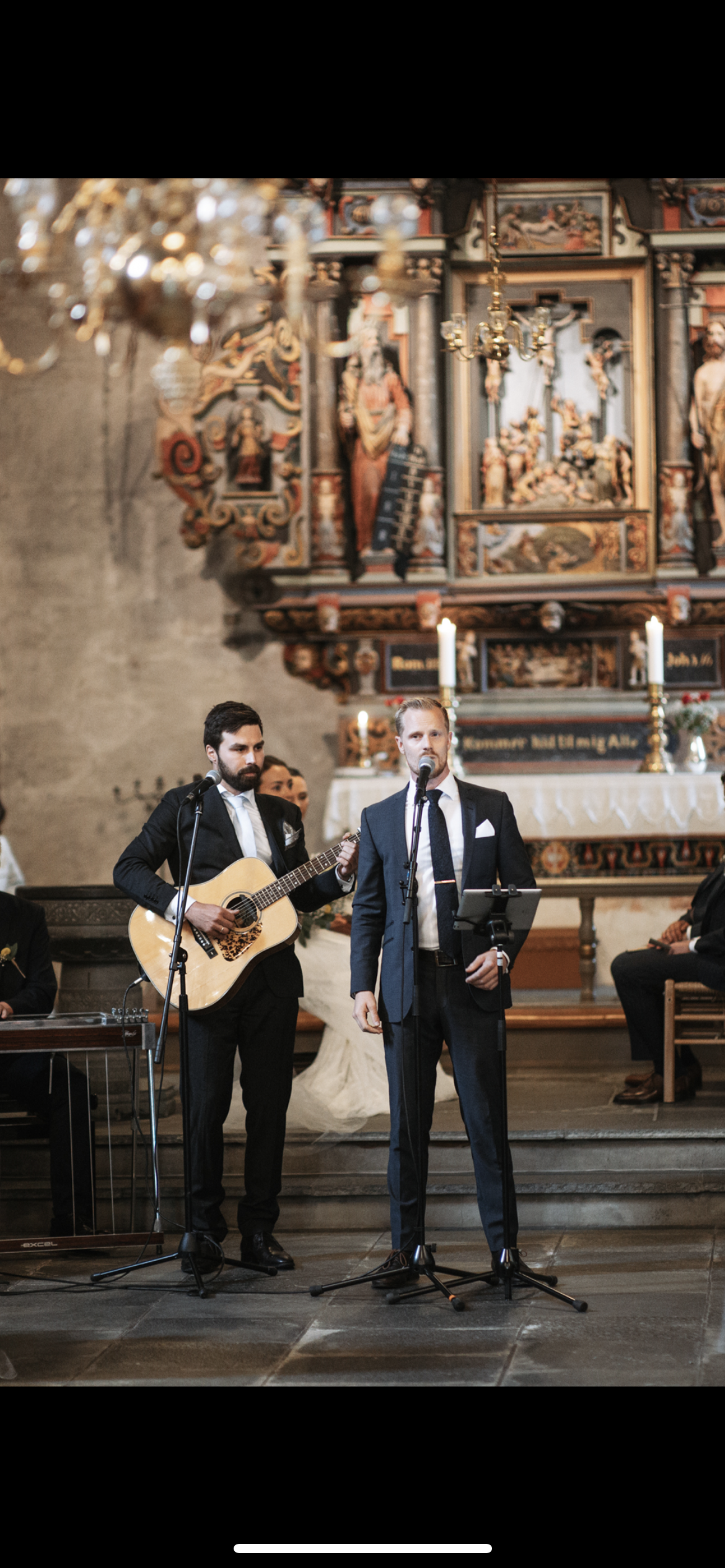 To menn står på en scene i en kirke, en med gitar og den andre med mikrofon. Bak dem er det et alter med stearinlys og religøse bilder.