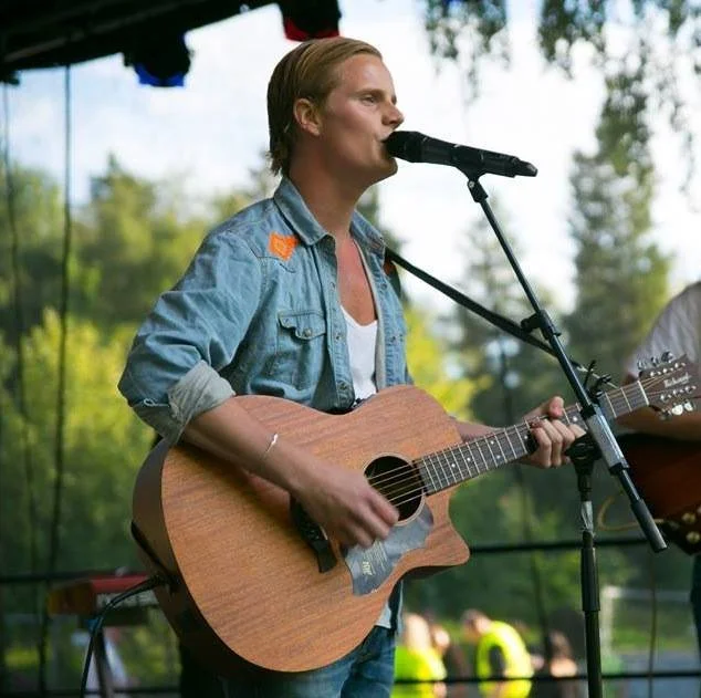 En person med kort blond hår som spiller gitar og synger innendørs mot et utendørs scenemiljø med trær i bakgrunnen.