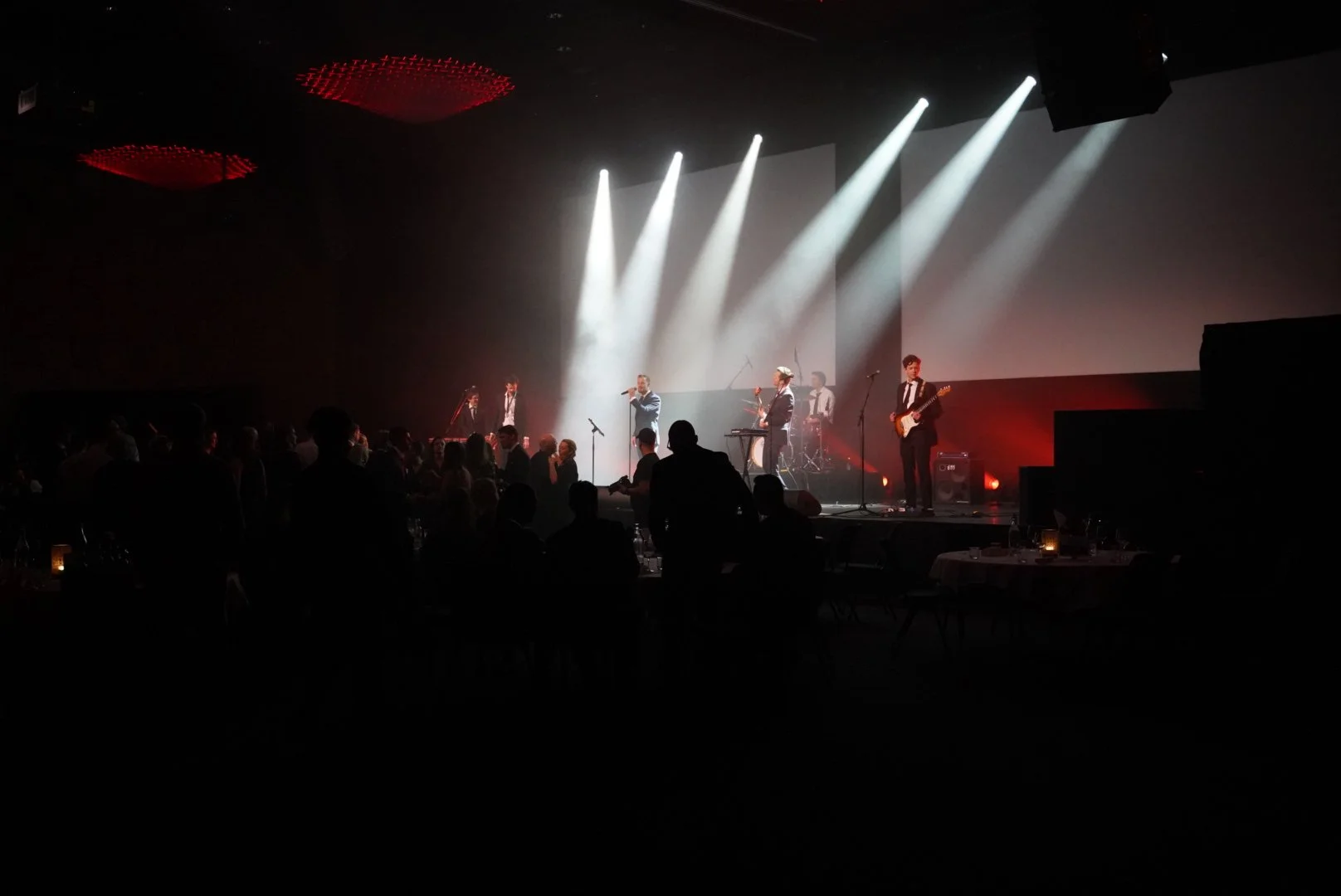 En kveldsscene med en live musikkopptreden på en scene med fem bandmedlemmer, inkludert en vokalist og gitarist, med et publikum som danser og nyter musikken. Scene belyses av hvite spotlights.