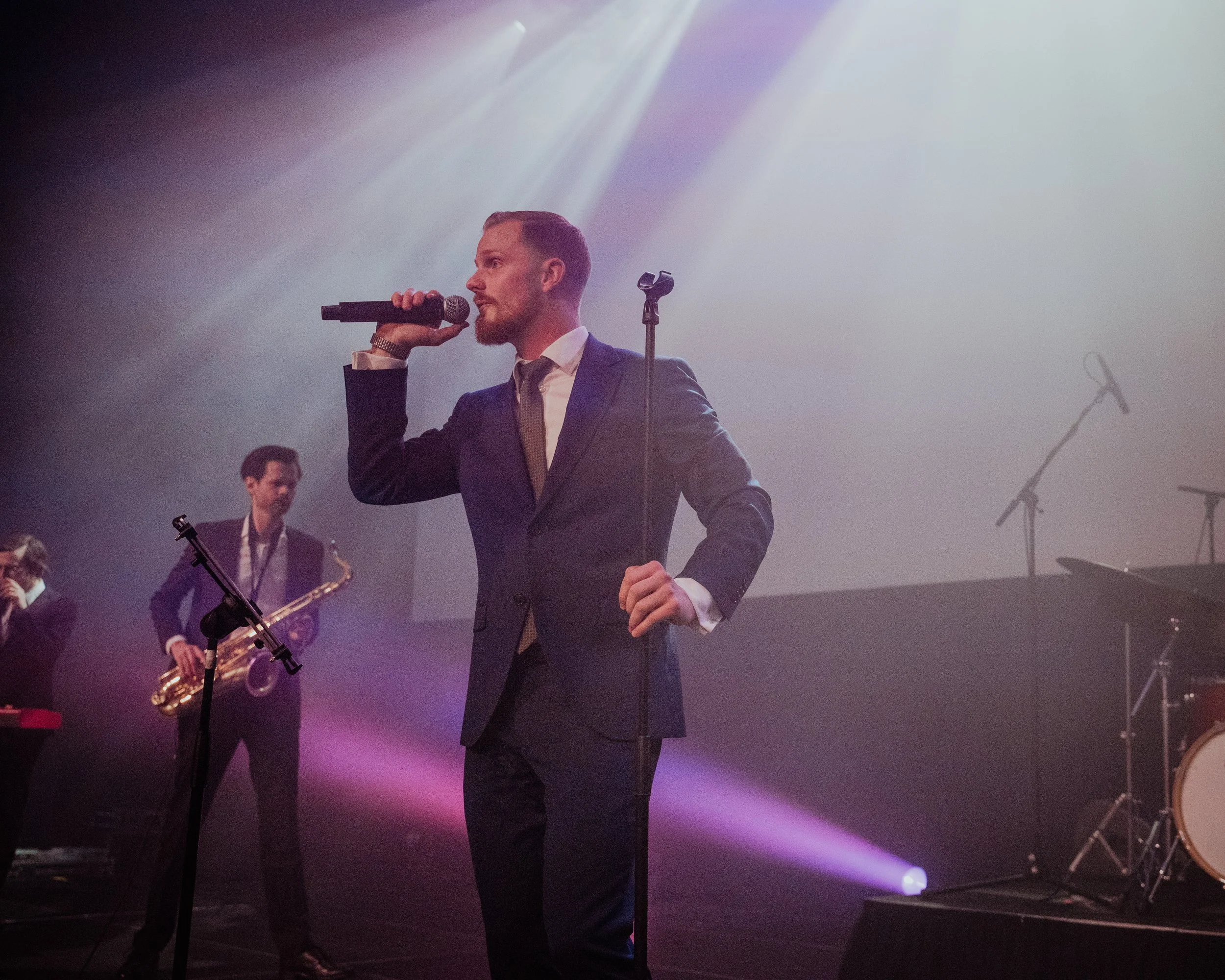 En mann i smal dress og slips holder mikrofon på en scene med andre musikere i bakgrunnen, og scenelys som skinner ned.