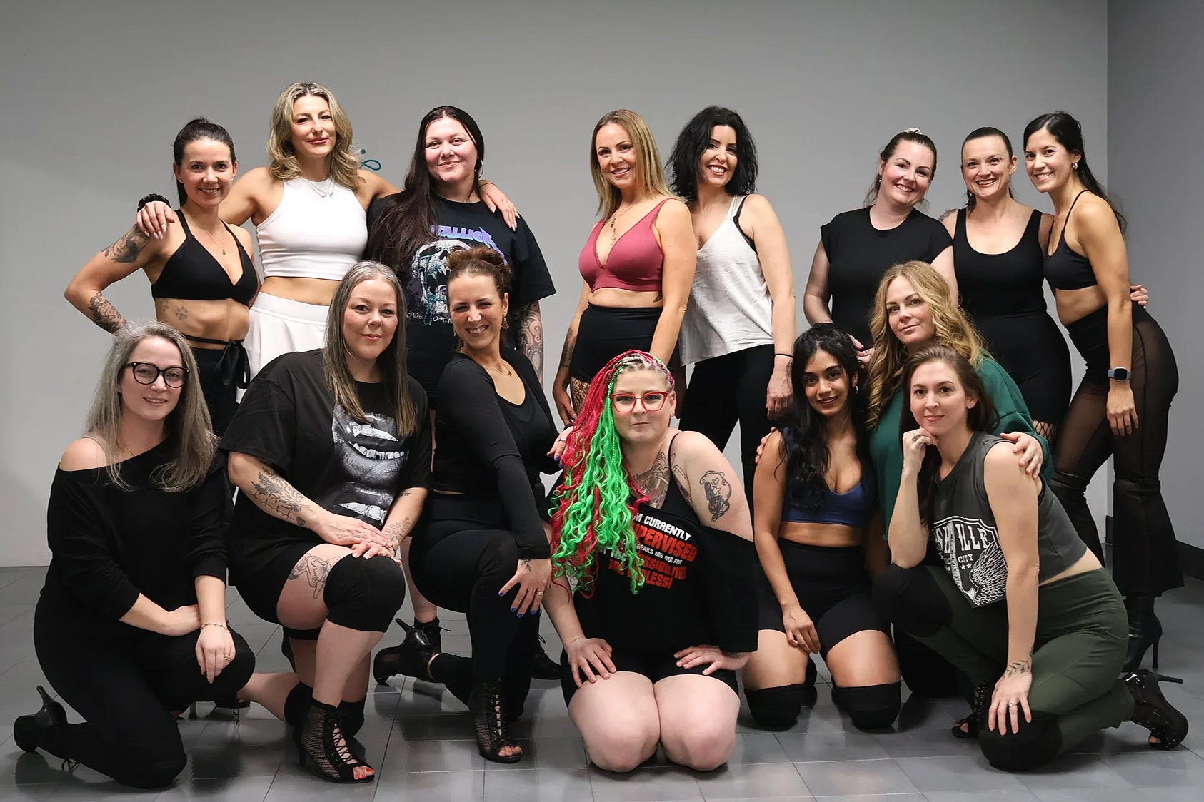 Group of 15 women posing together in front of a gray wall, some standing, some kneeling or sitting, wearing casual and activewear clothing.
