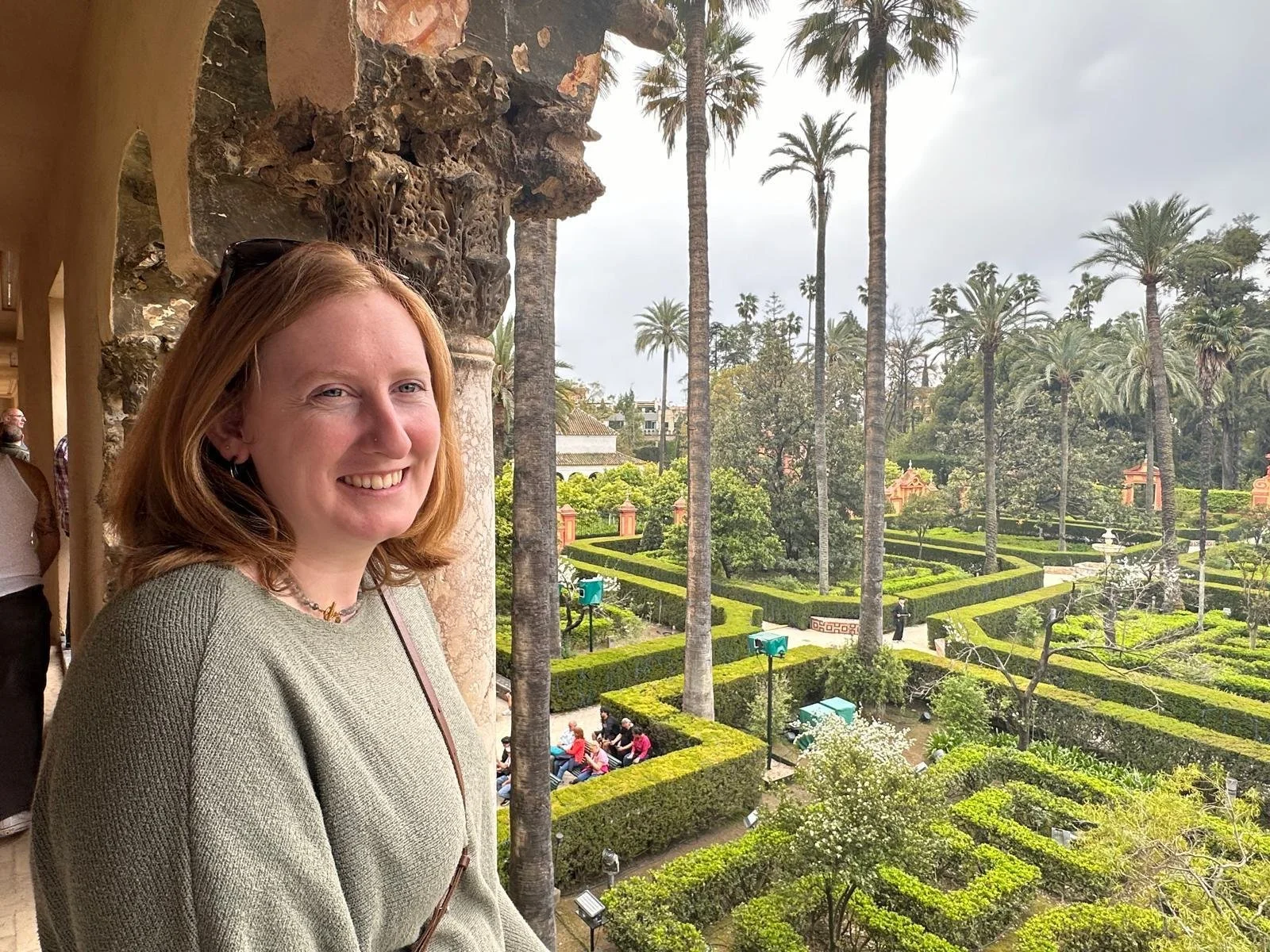 A young woman with red hair smiling while leaning against a stone column, overlooking a lush, well-manicured garden with tall palm trees and a fountain, on a cloudy day.
