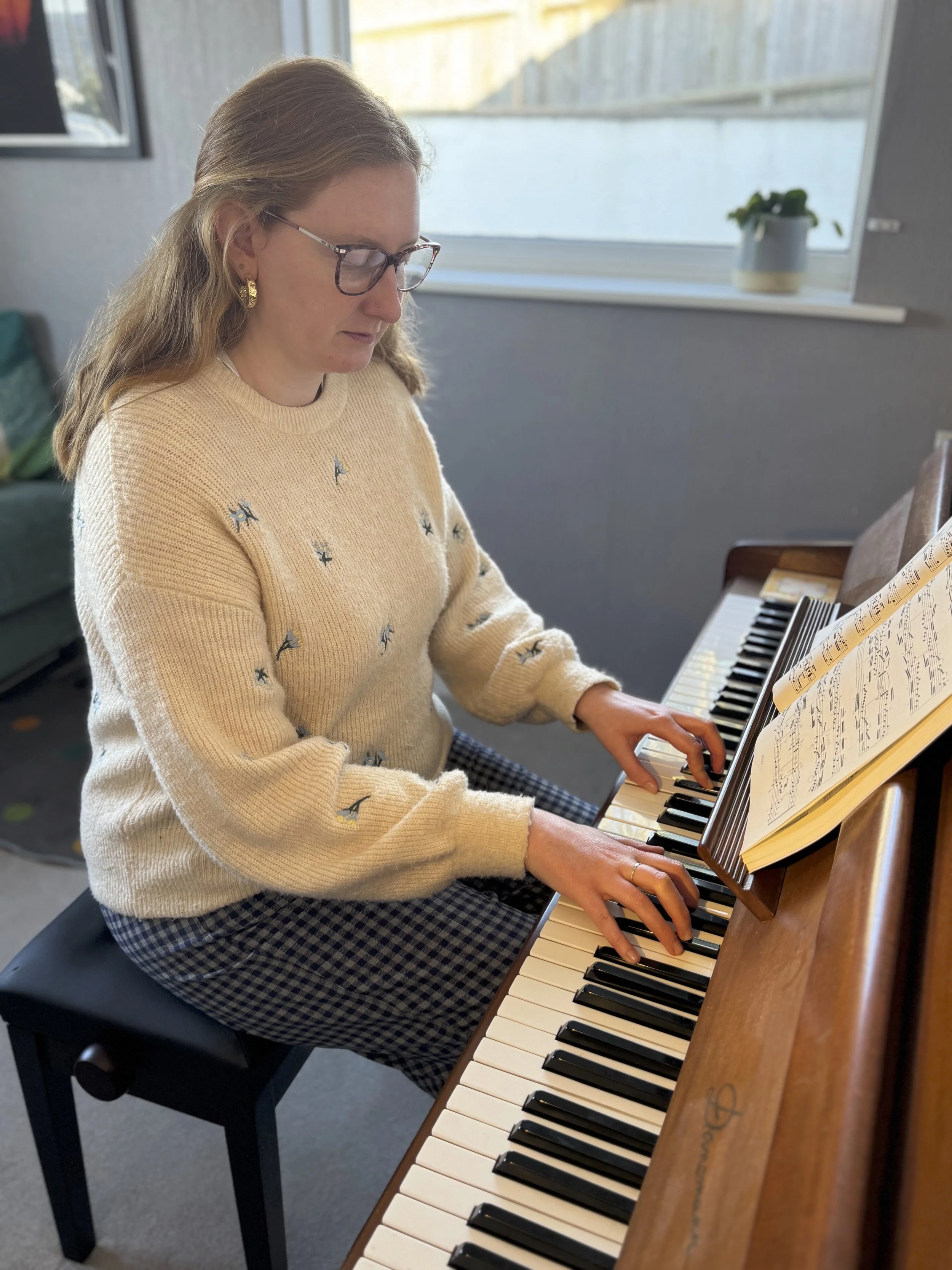 A woman with glasses and long hair playing the piano, looking at sheet music.
