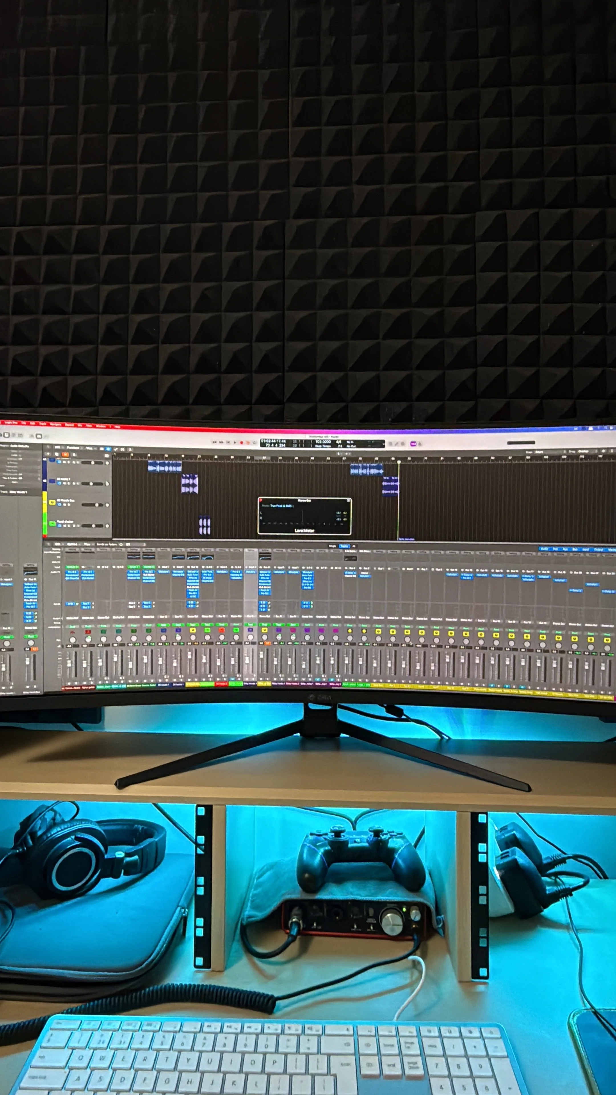 Computer monitor displaying music production software in a home studio with soundproof foam panels on the wall, a keyboard, headphones, gaming controllers, and audio equipment on a desk.
