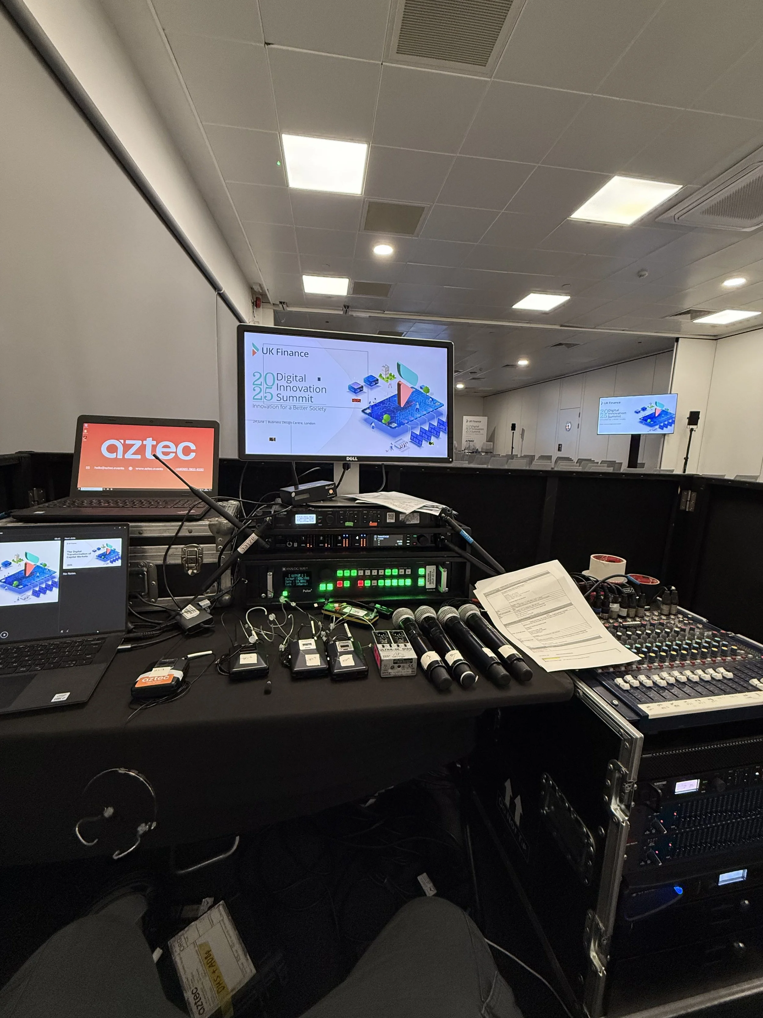View of a conference room setup with a focus on audio and visual equipment, including microphones, a sound mixer, multiple monitors displaying a presentation titled 'UK Finance Digital Innovation Summit 2025,' and a laptop with 'aztec' branding.