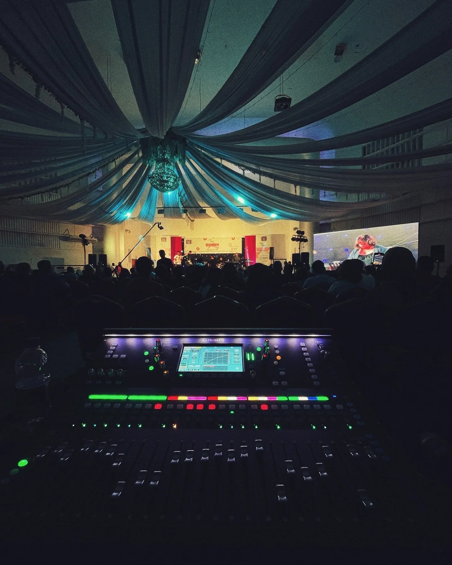 View of a stage with a live band performing under blue lighting, decorated with draped fabric and a chandelier. Audience silhouettes and a large screen displaying a performer are visible. In the foreground, a DJ mixing console with colorful illuminat